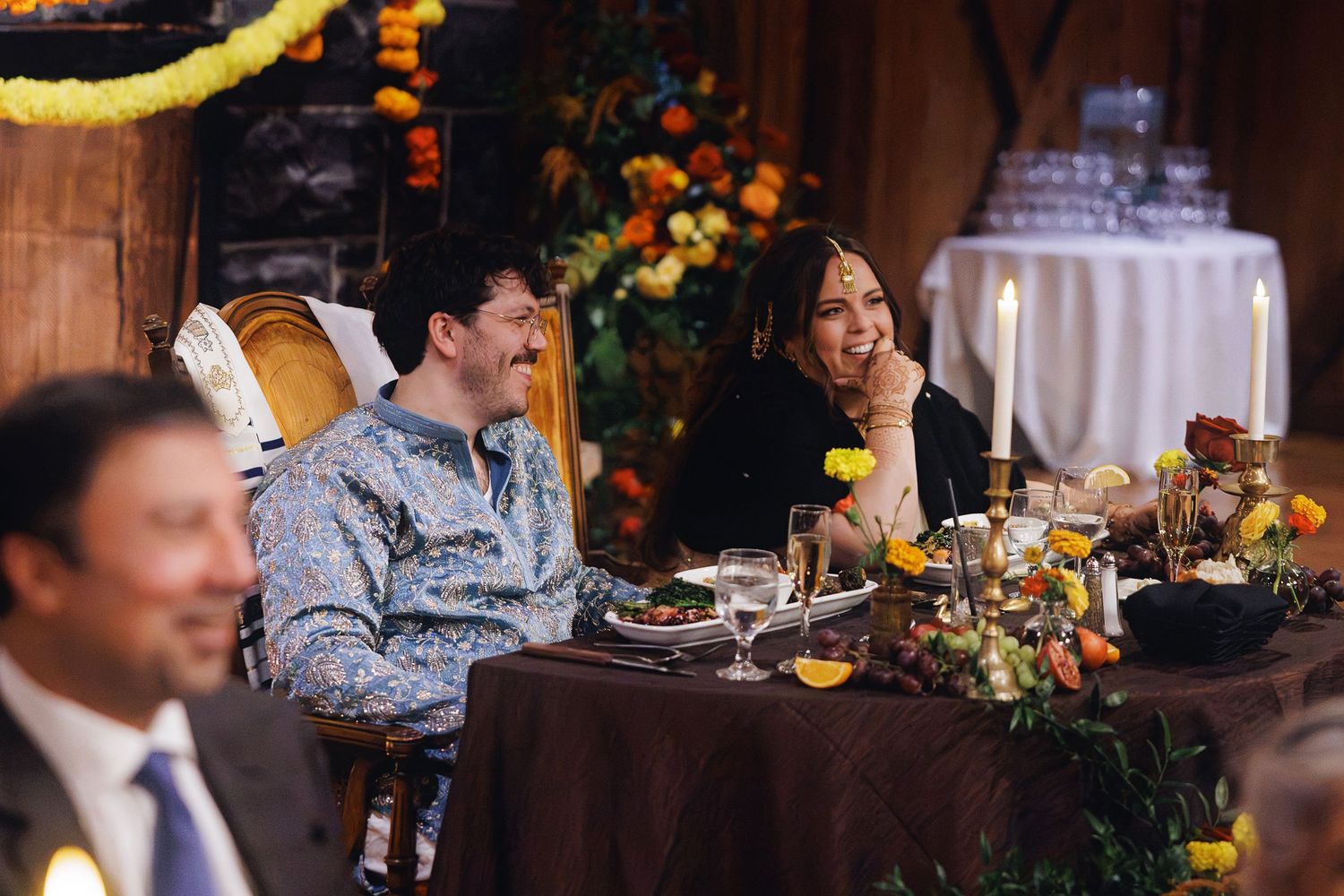 Two guests laugh joyfully at a decorated dinner table with candles and floral arrangements at an elegant event.