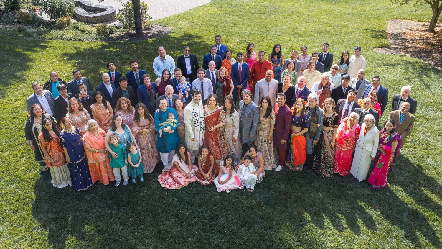 Large diverse wedding group photo outdoors on green lawn, guests in colorful traditional Indian attire and formal wear.