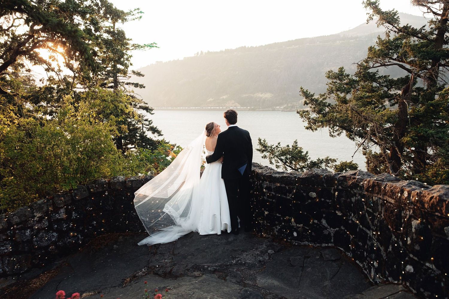 Bride and groom stand at stone overlook gazing at scenic lake and mountain views during outdoor wedding ceremony.