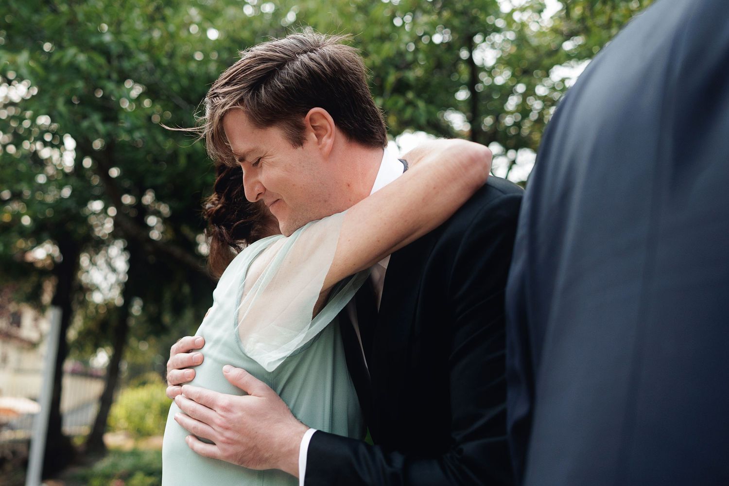 A man in a dark suit tenderly embraces a woman in a mint green dress outdoors among green trees.