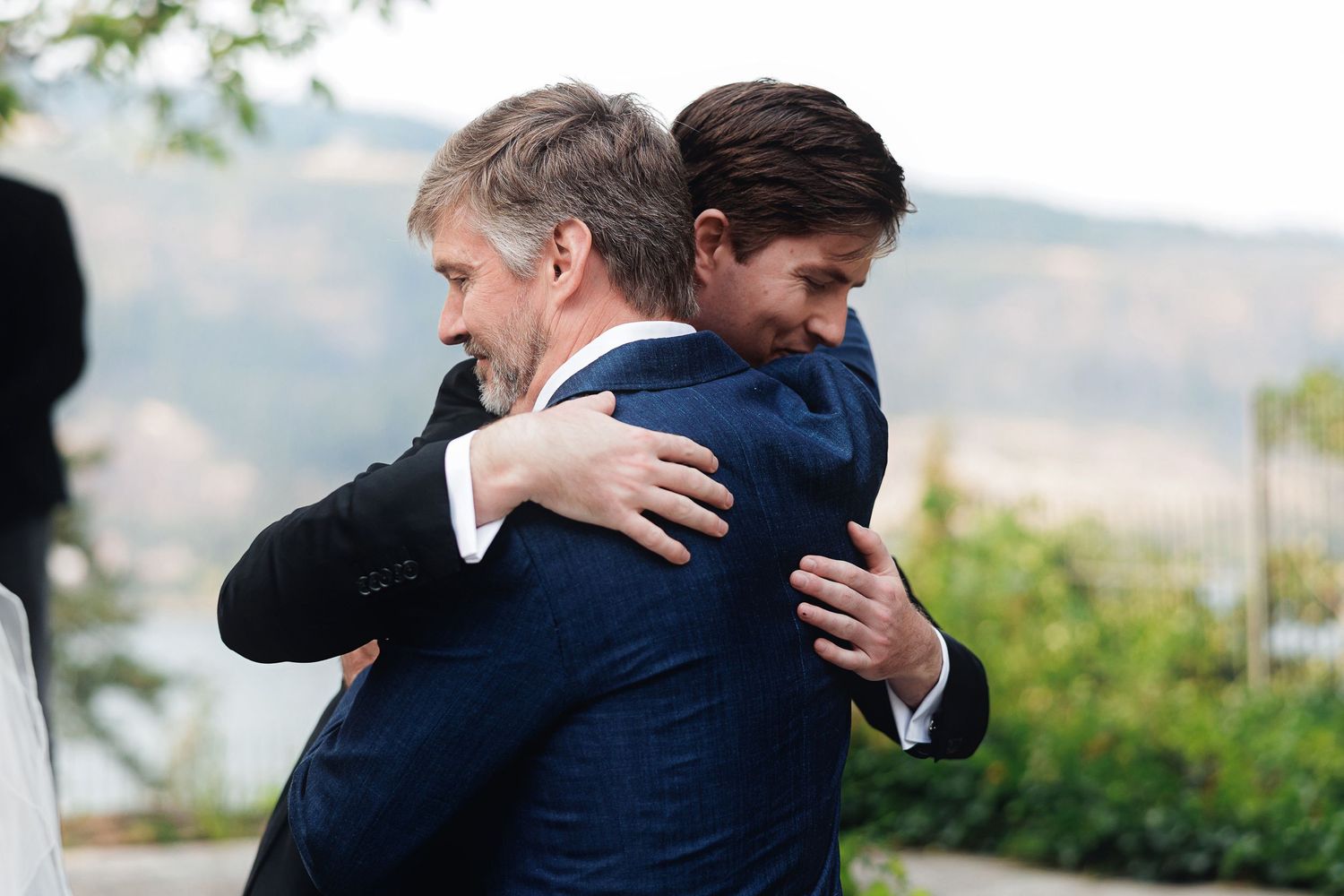 Two men in navy suits share a warm embrace outdoors with a scenic mountain landscape in the background.