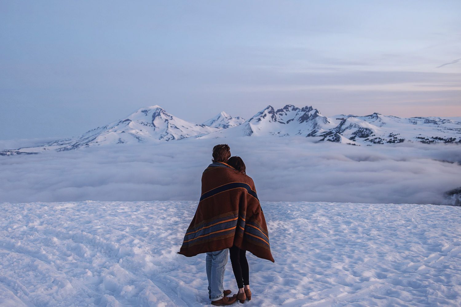 A person wrapped in a blanket stands on a snowy mountaintop overlooking a sea of clouds and distant peaks at dusk.