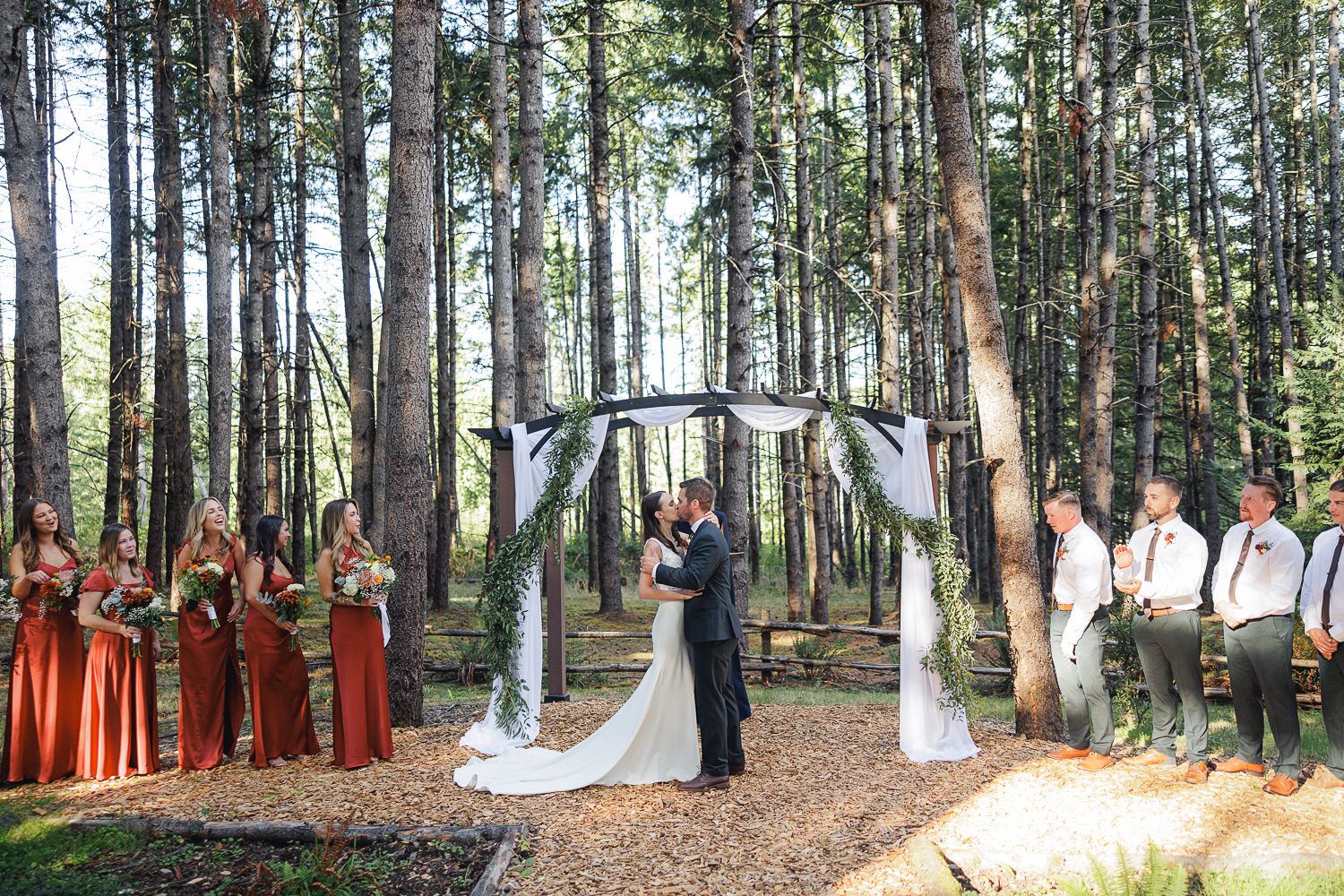 Forest wedding ceremony with bridal party under rustic arch surrounded by tall pine trees.