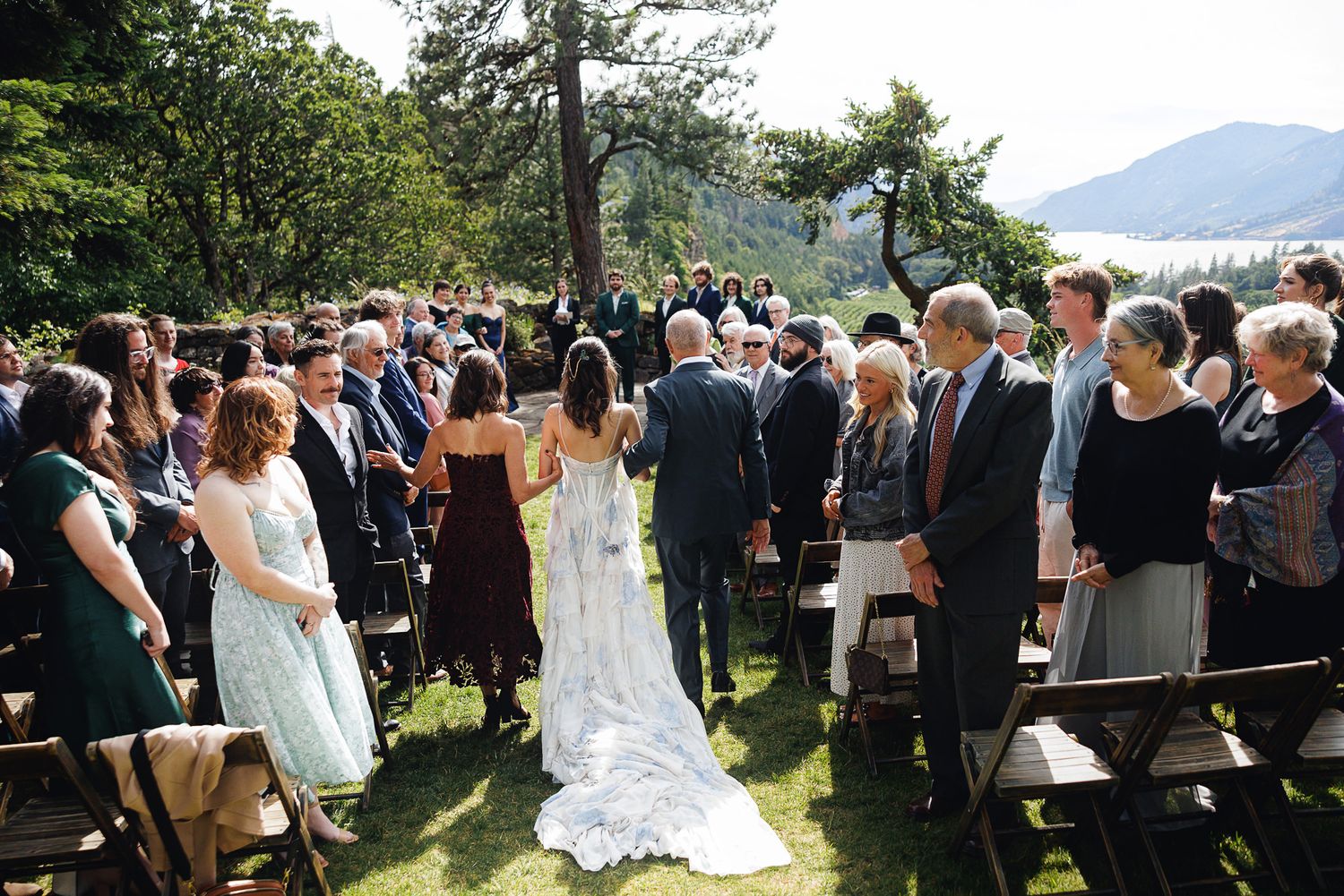 Bride and groom walk down outdoor wedding aisle surrounded by guests, with scenic mountain and lake views in background.