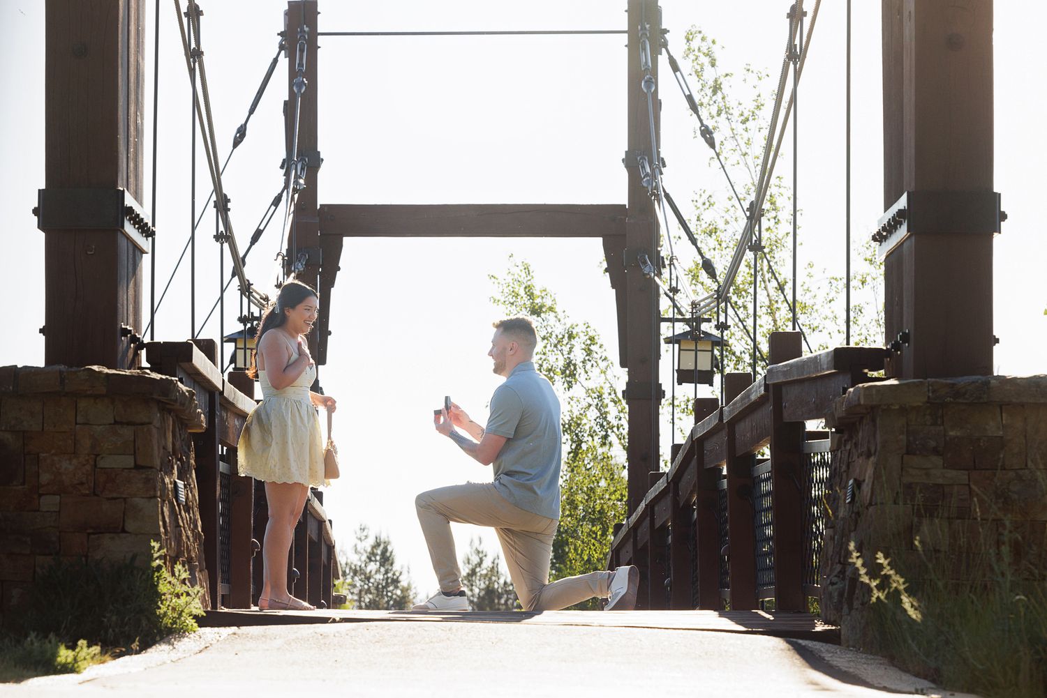 Man proposing to woman on a rustic suspension bridge outdoors on a sunny day.
