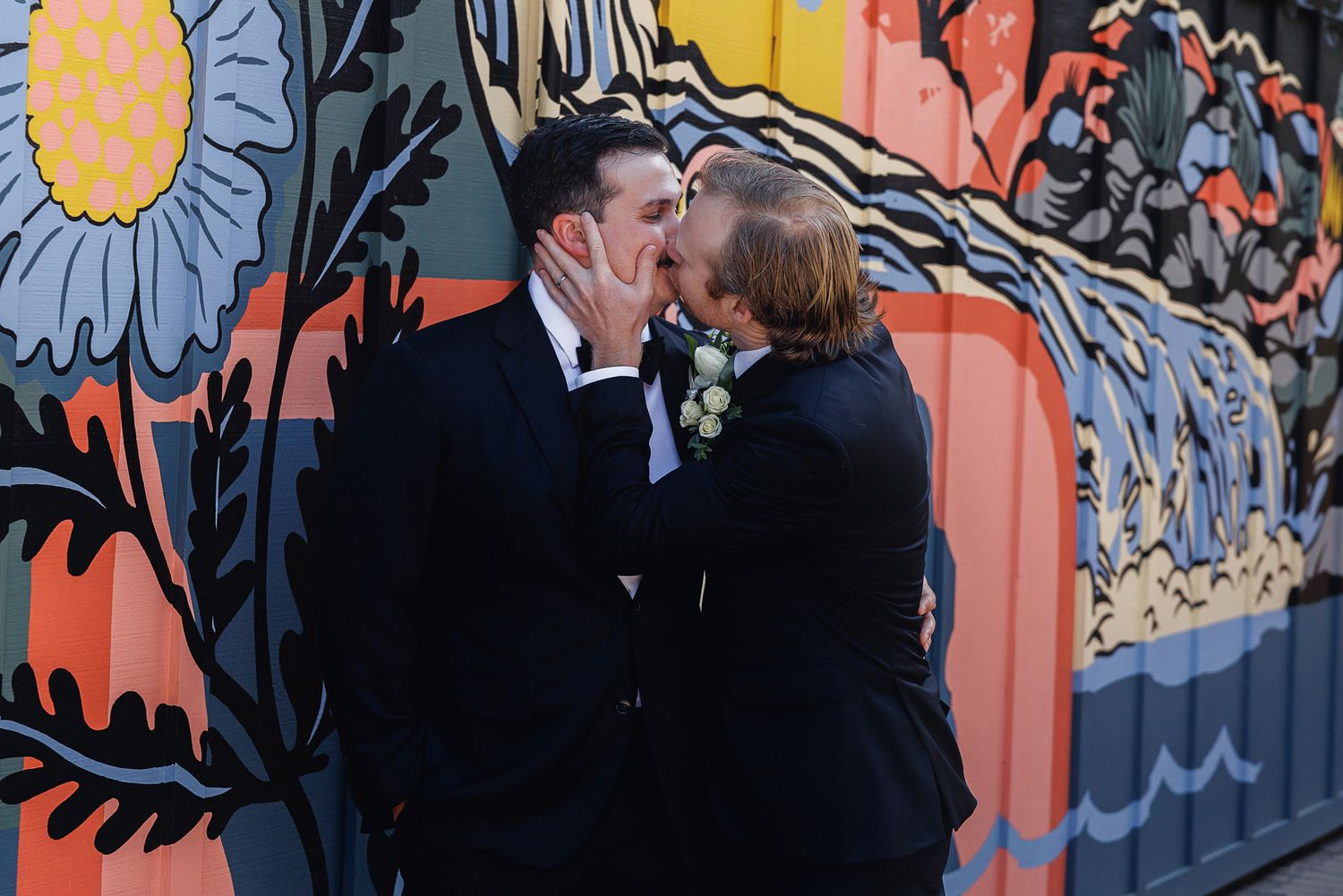 Two grooms share a kiss in front of a colorful floral mural on their wedding day.