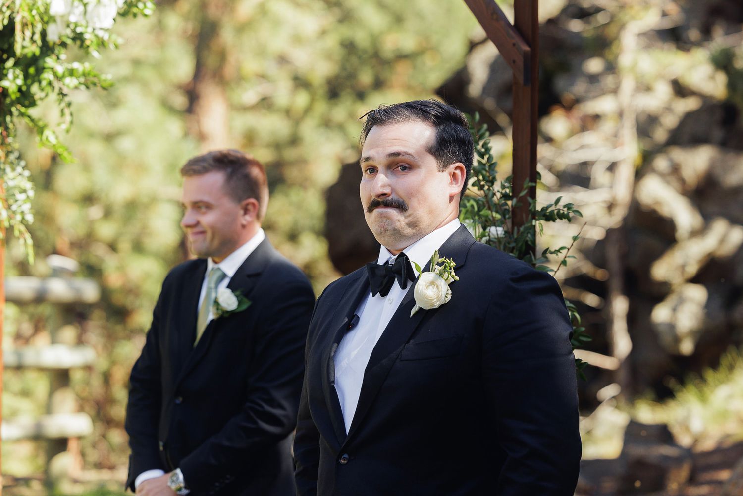 Groom in black tuxedo with boutonniere stands at outdoor wedding altar, with groomsman in background.