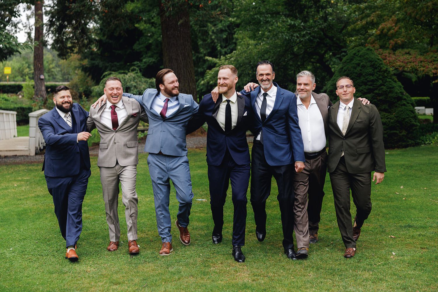 Seven men in suits laughing and posing together on green lawn at outdoor wedding celebration.
