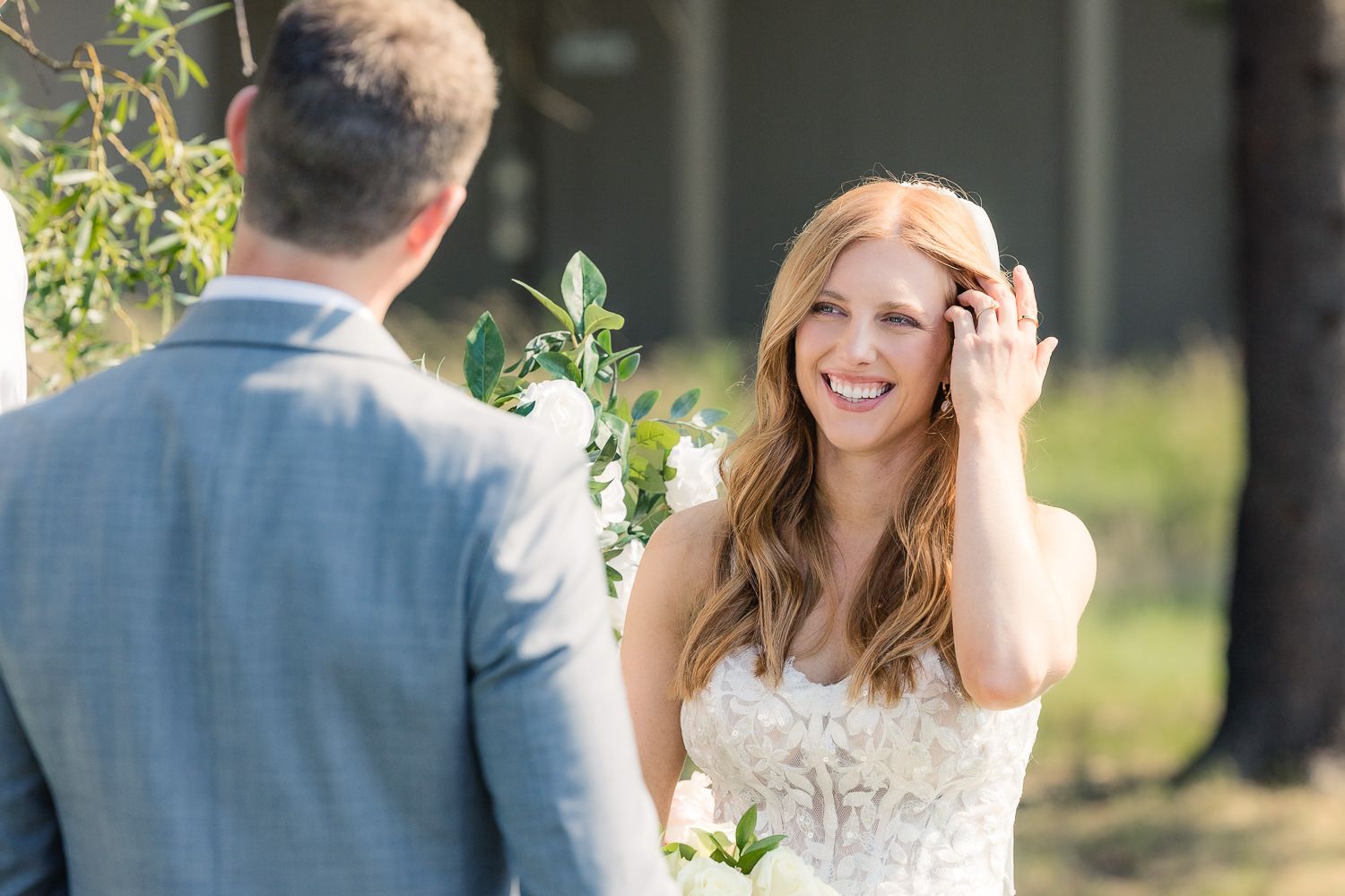 A beaming bride in a white lace dress shares a candid moment during an outdoor wedding ceremony.