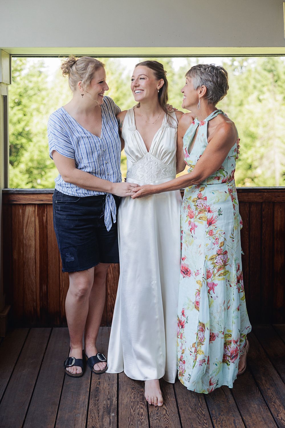 A bride is hugged by her sister and mother standing together on a wooden porch in a candid moment