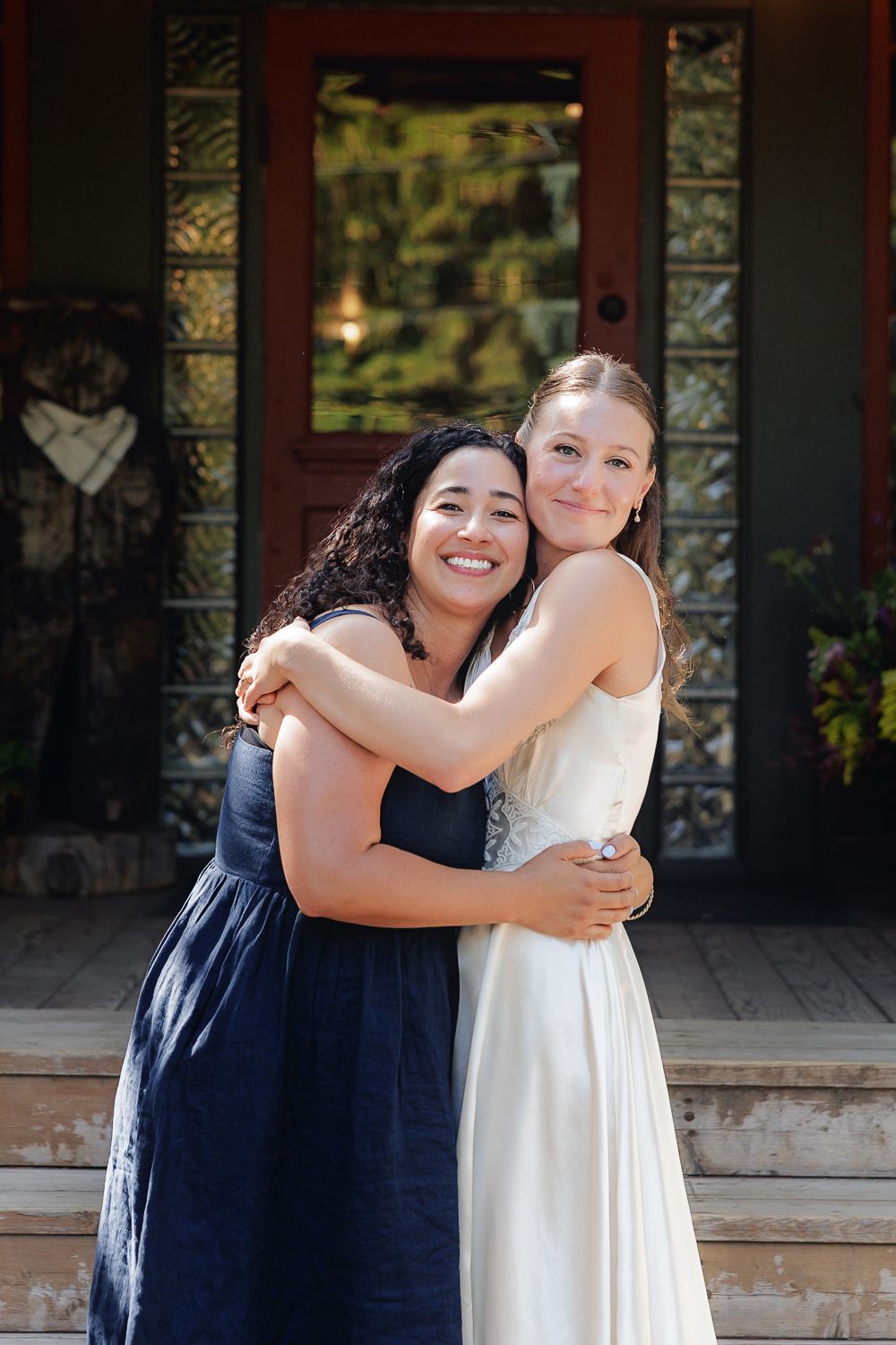 Two friends embrace and smile while standing on wooden steps outside a building with decorative windows.