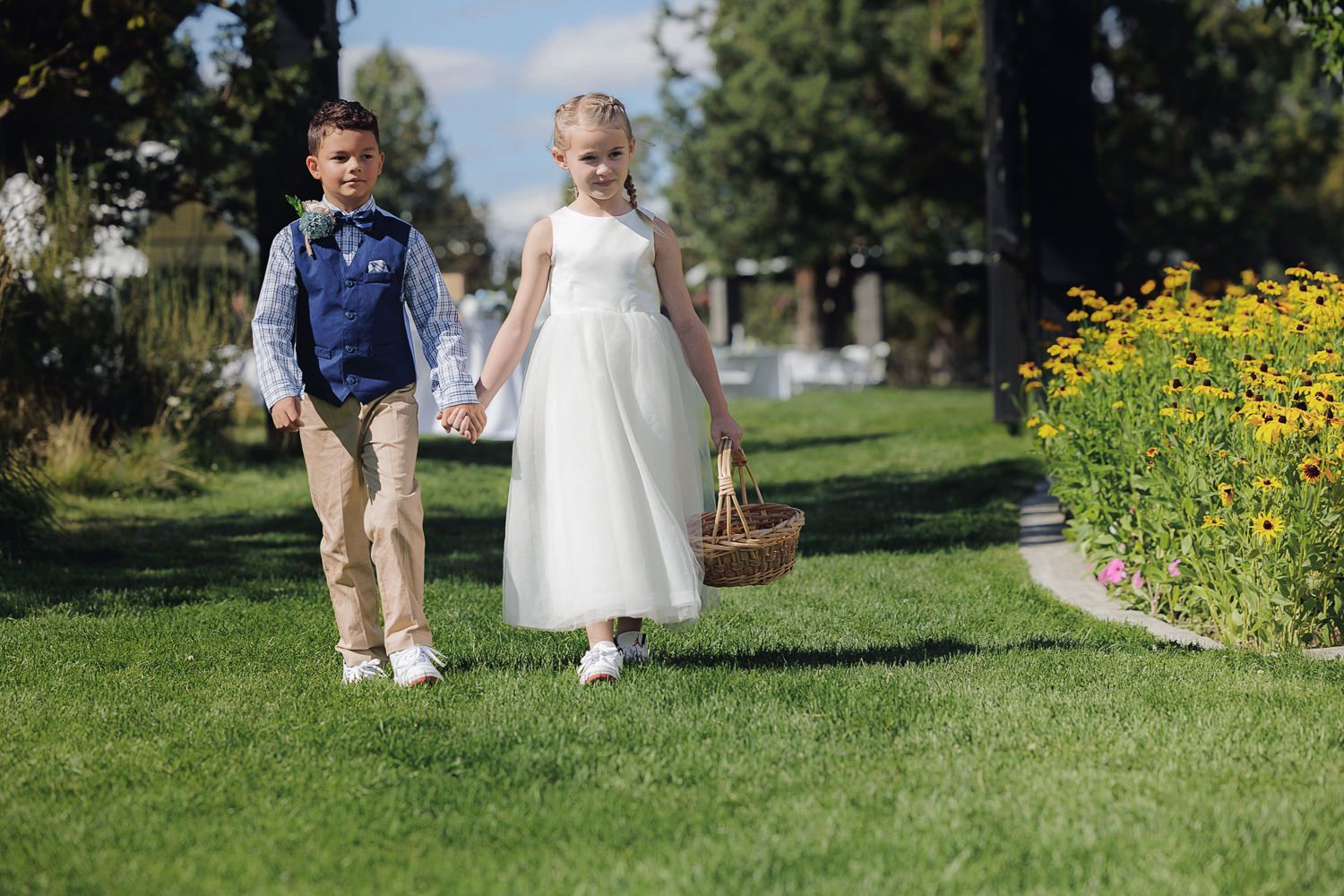 Two children walk hand in hand through a sunny garden path bordered by bright yellow wildflowers.