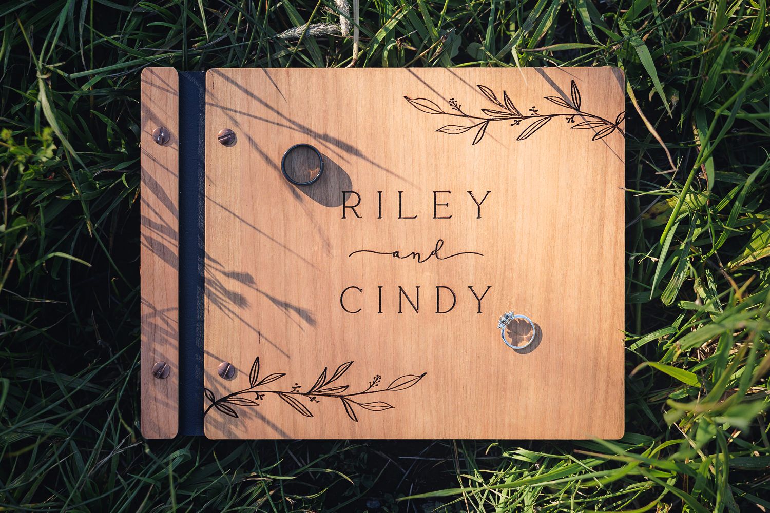 Wooden photo album cover with names Riley and Cindy engraved alongside decorative leaf designs resting on grass.