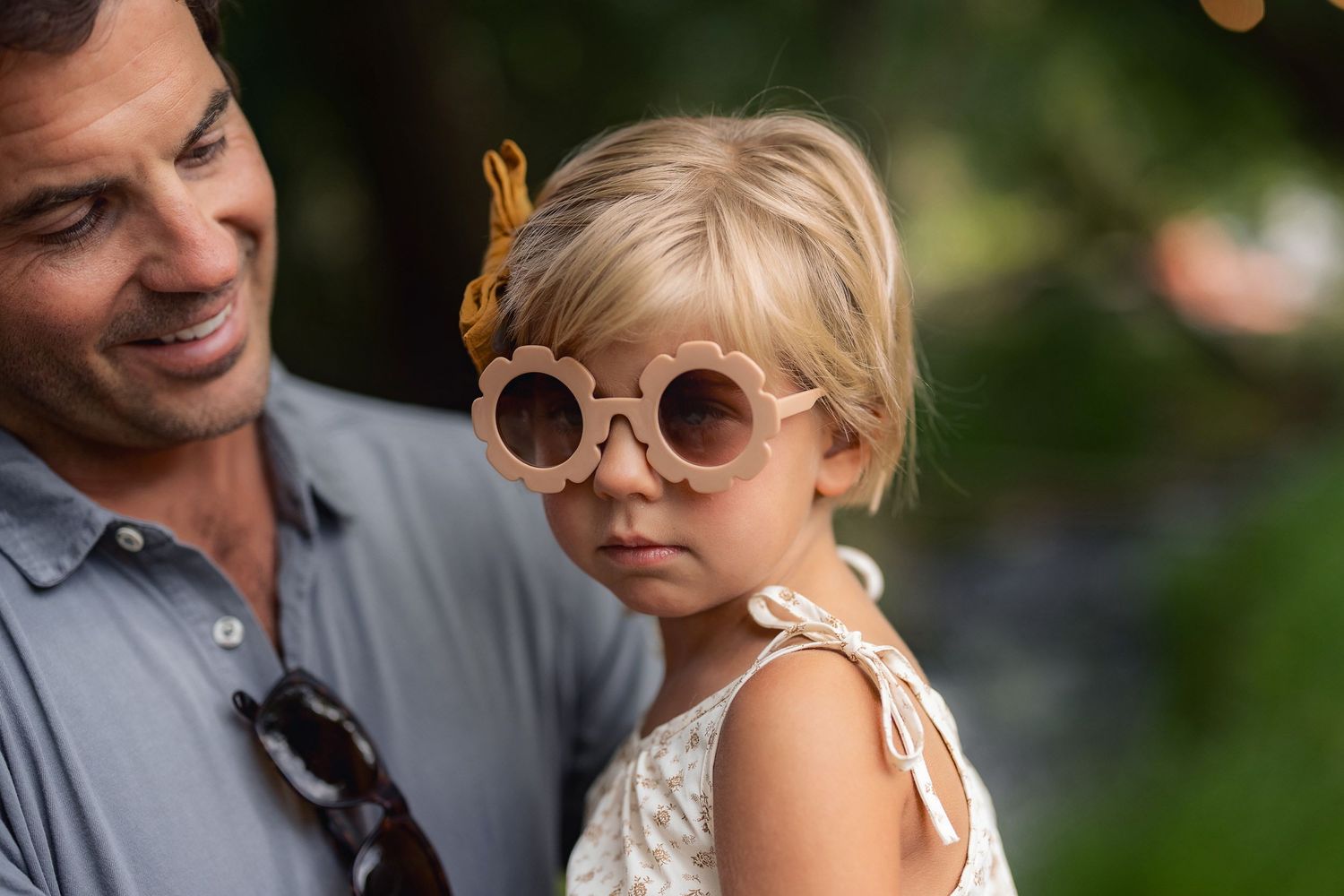 A small child wearing round sunglasses and a white dress smiles while being held by an adult in a gray shirt.