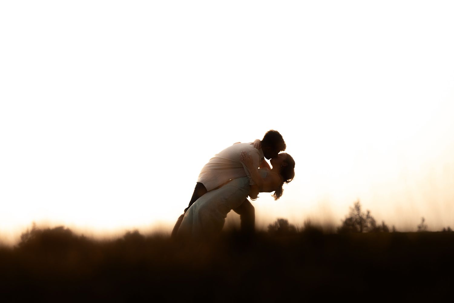 Romantic silhouette of a couple sharing a tender kiss at sunset in an open field.