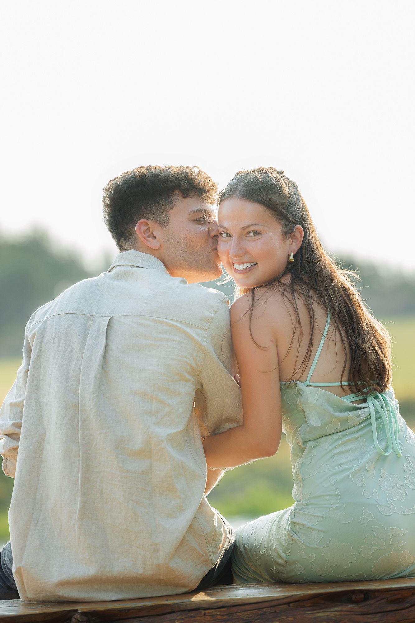 A young couple sitting outdoors, man kissing woman on cheek as she smiles at camera in golden light.