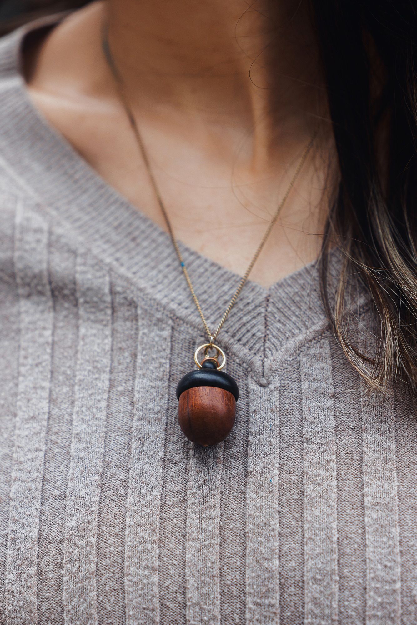 Close-up of a wooden acorn pendant necklace on a delicate gold chain against a gray ribbed sweater.