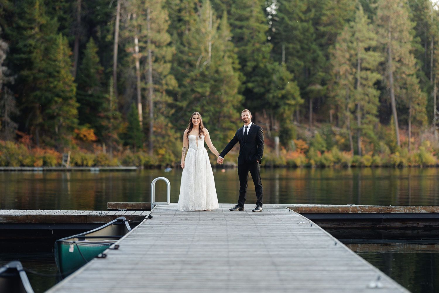 Bride and groom hold hands on a wooden dock surrounded by a serene lake and autumn forest backdrop.