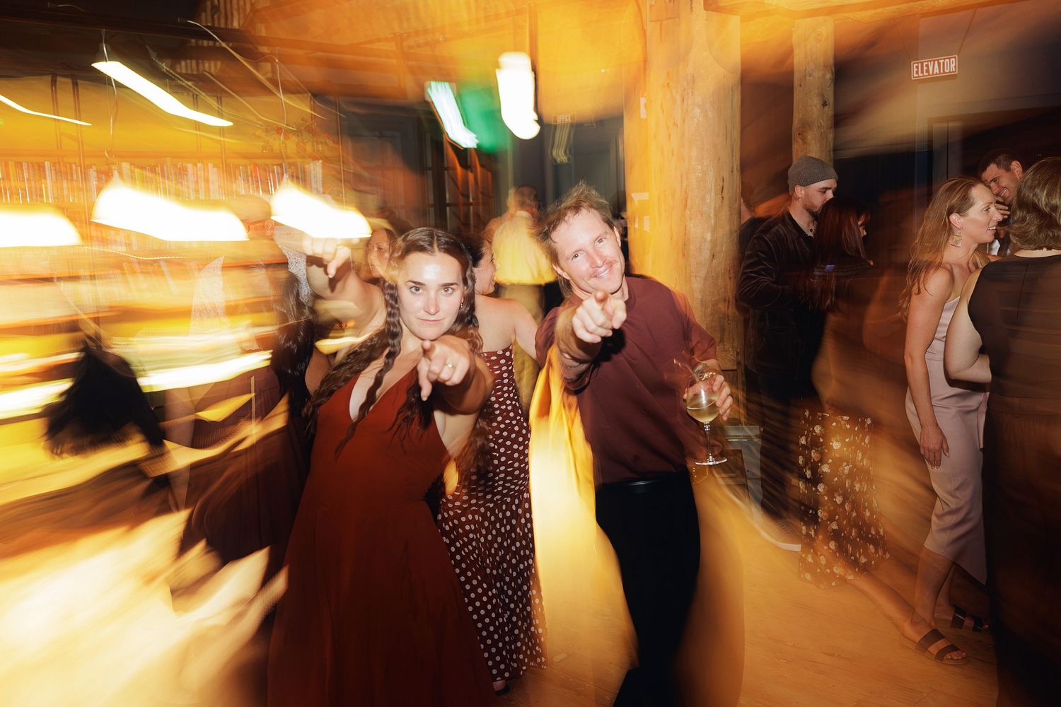 Two people dancing and pointing at camera at a lively party with warm golden lighting and motion blur effect.