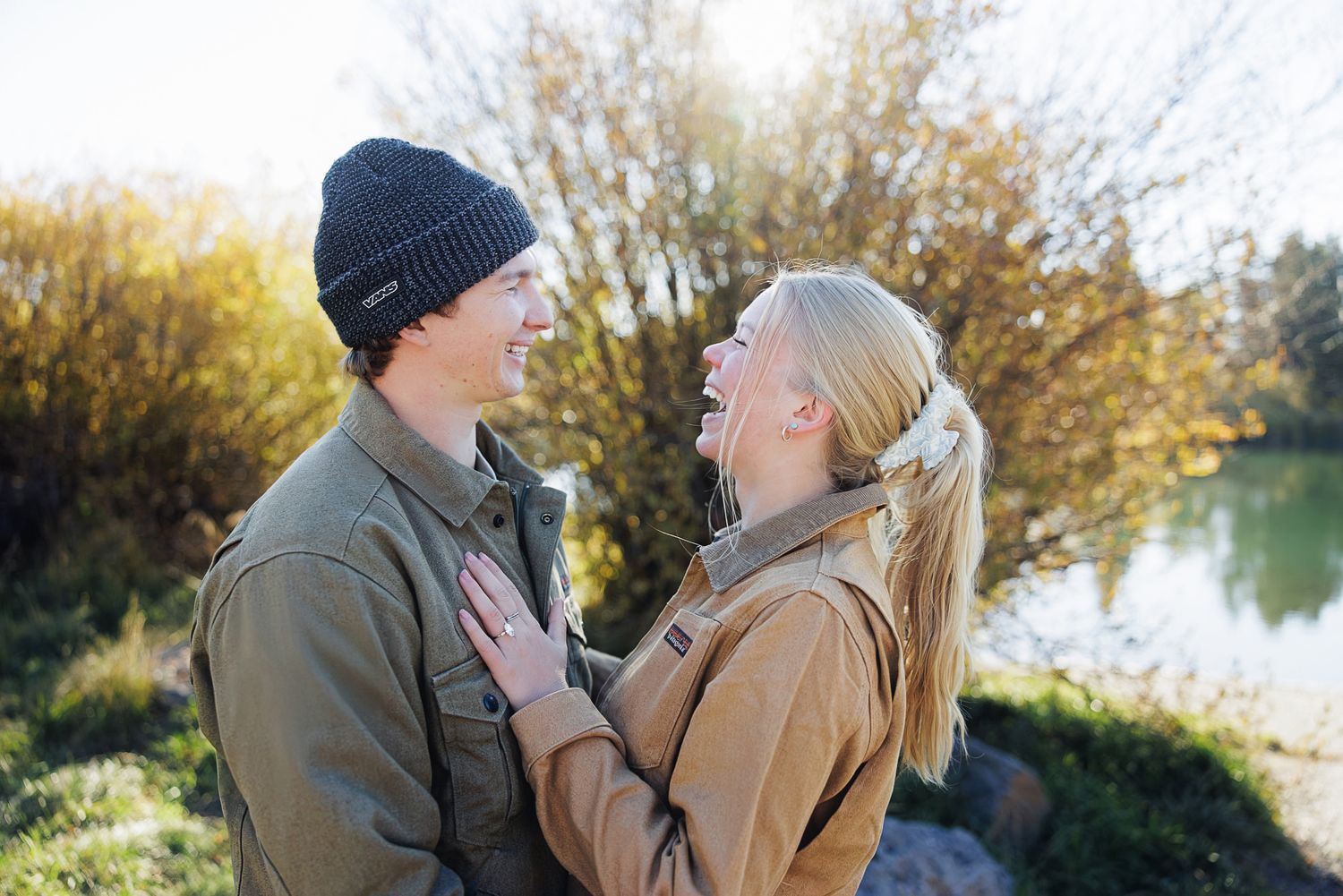 A smiling couple in casual jackets laughs together outdoors by a calm lake, surrounded by golden autumn foliage.
