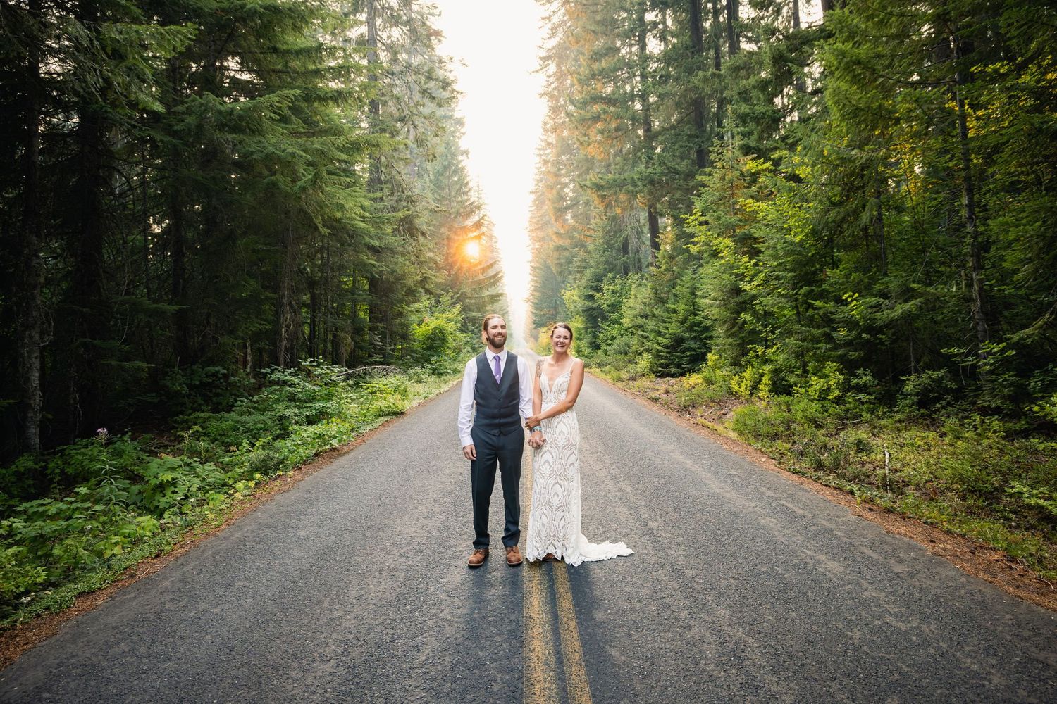 A couple in wedding attire standing together on a forest road during sunset.