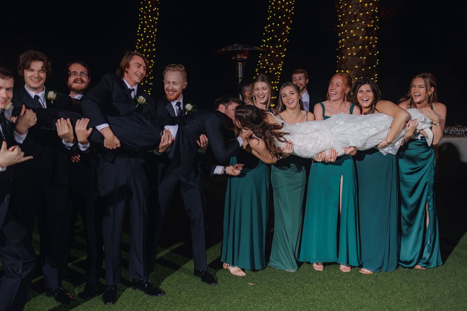 Wedding party in navy suits and emerald dresses poses playfully while holding someone horizontally outdoors.