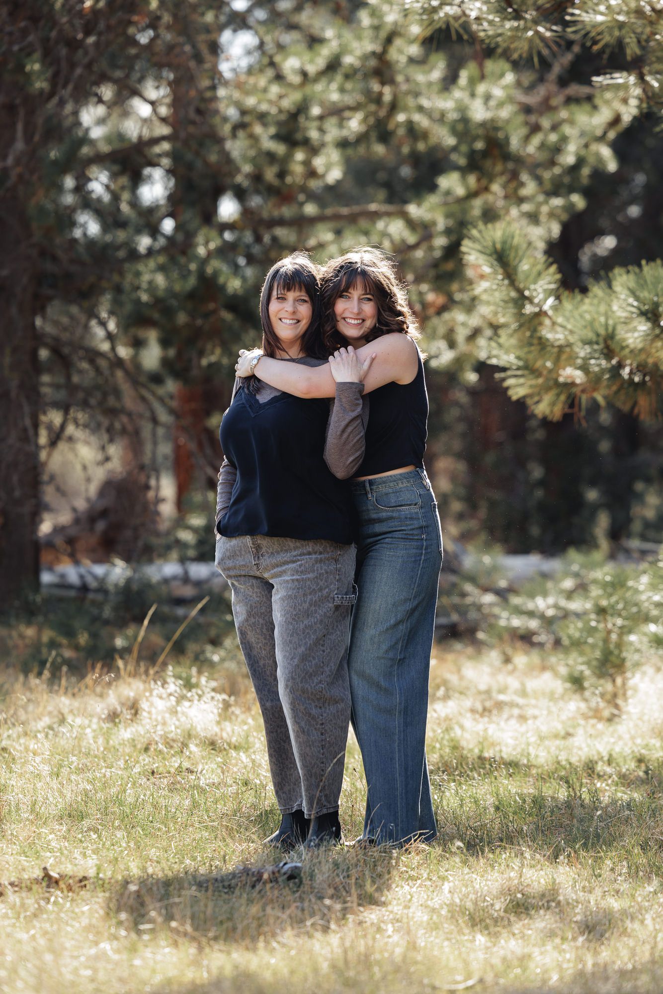 Two women hugging and smiling outdoors among pine trees in a sunny forest meadow.