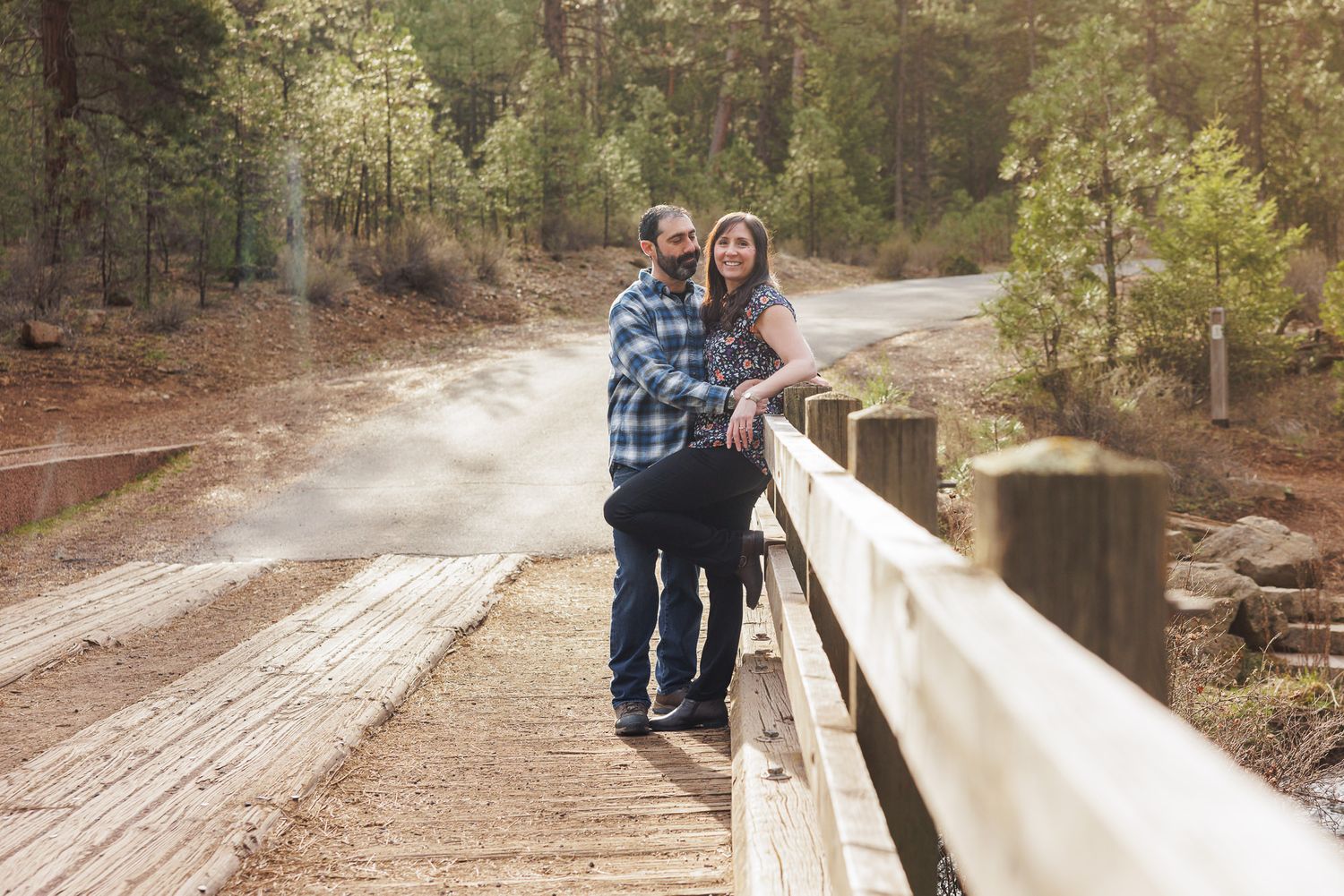 Couple leaning on wooden fence bridge surrounded by pine forest on a scenic dirt road.