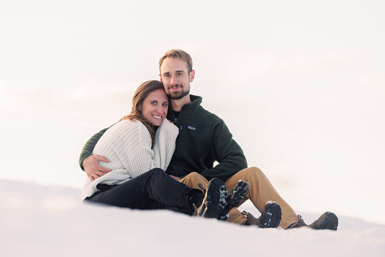 Couple sitting together on snow, smiling, wearing winter clothes including a white sweater and green jacket.