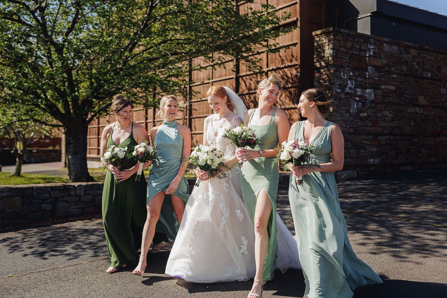 Bride in white gown walks with four bridesmaids in sage green dresses, holding white bouquets outside a brick building.