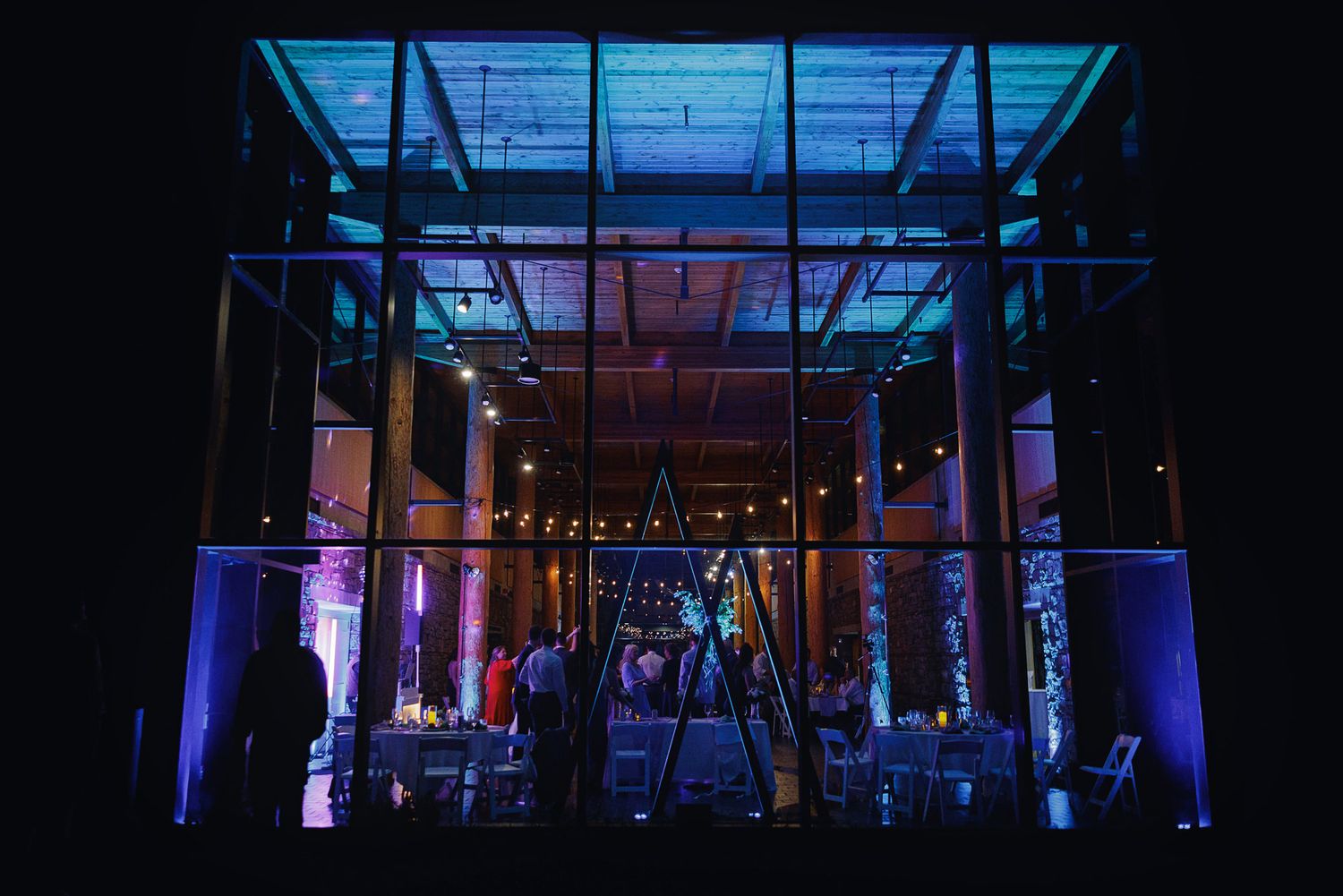 Elegant event venue glowing with blue and purple lights, featuring glass walls and string lights inside.