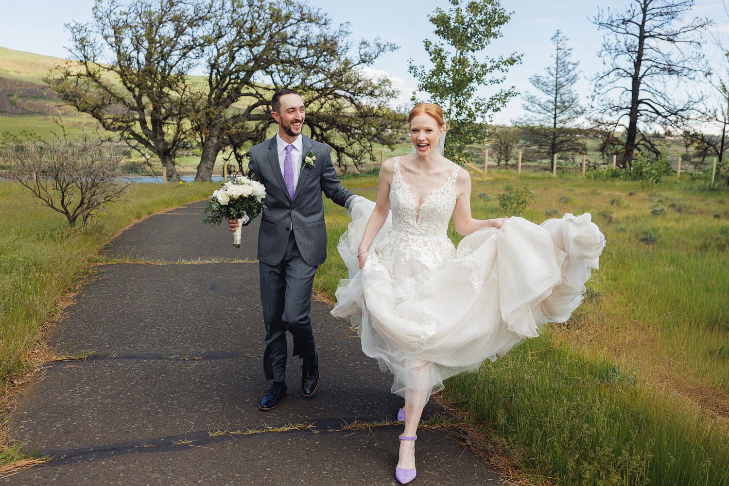 A bride in a white gown and groom in a gray suit walk hand in hand down a scenic path surrounded by green hills.