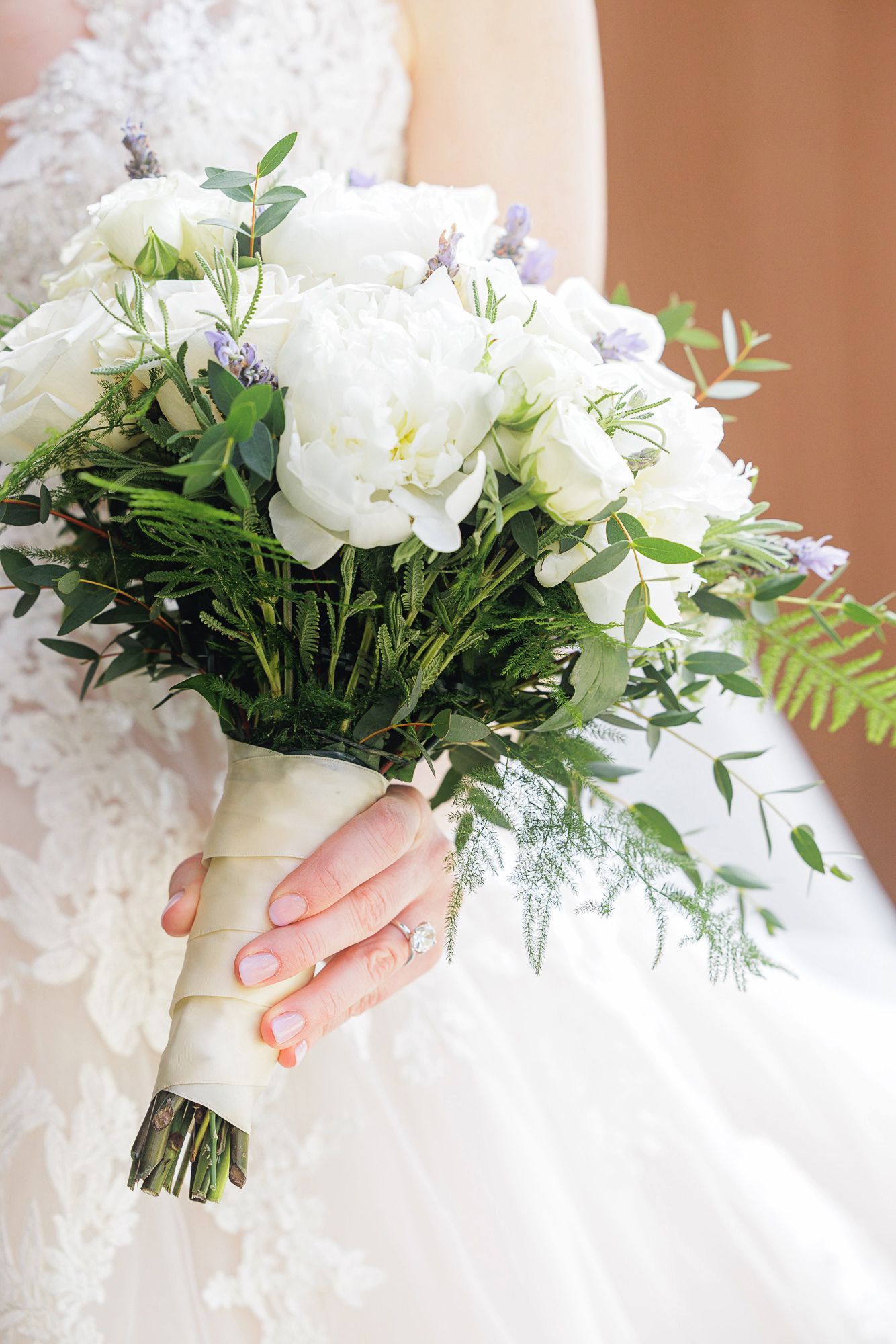 Bride holding a lush white peony and greenery wedding bouquet wrapped in white ribbon, wearing a lace gown.