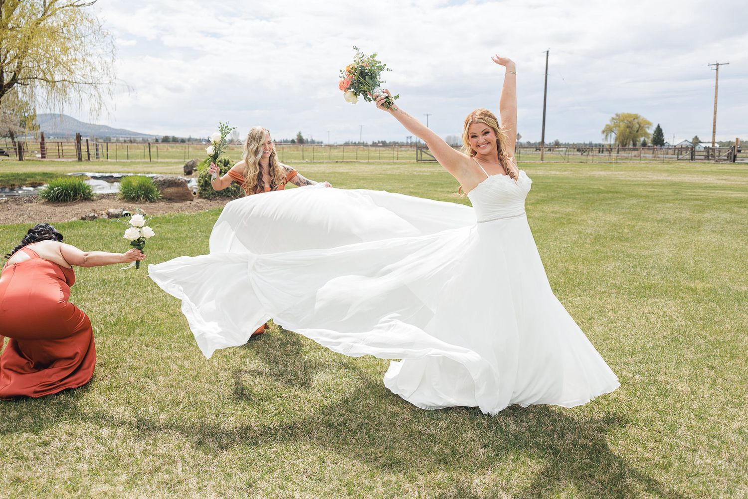 Joyful bride in white gown twirling outdoors while bridesmaids assist with her flowing dress on a sunny wedding day.