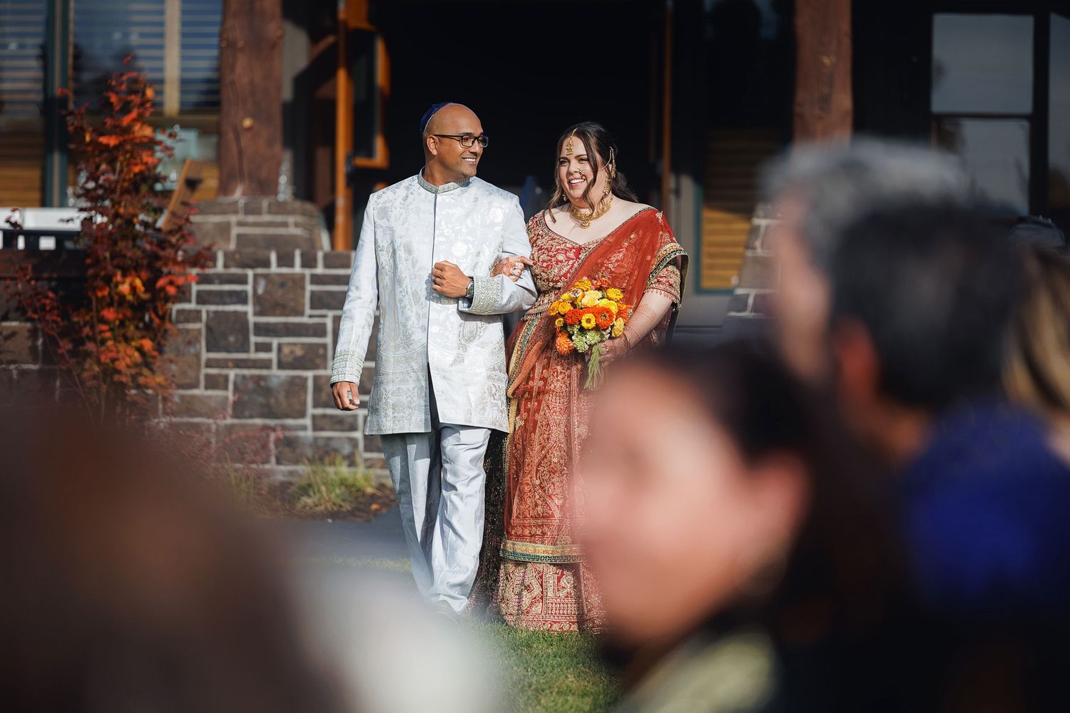 Couple walks down aisle in Indian wedding attire, bride in red saree with flowers, groom in white sherwani, outdoor ceremony.
