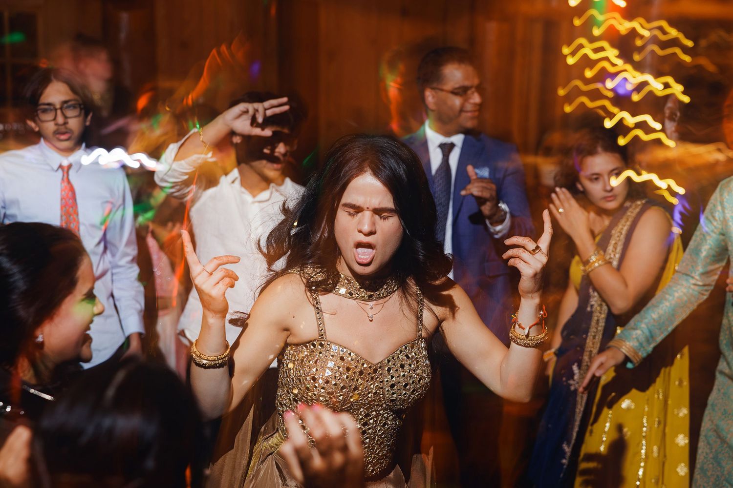 Woman in gold dress dancing energetically at a vibrant Indian wedding reception with guests celebrating around her.