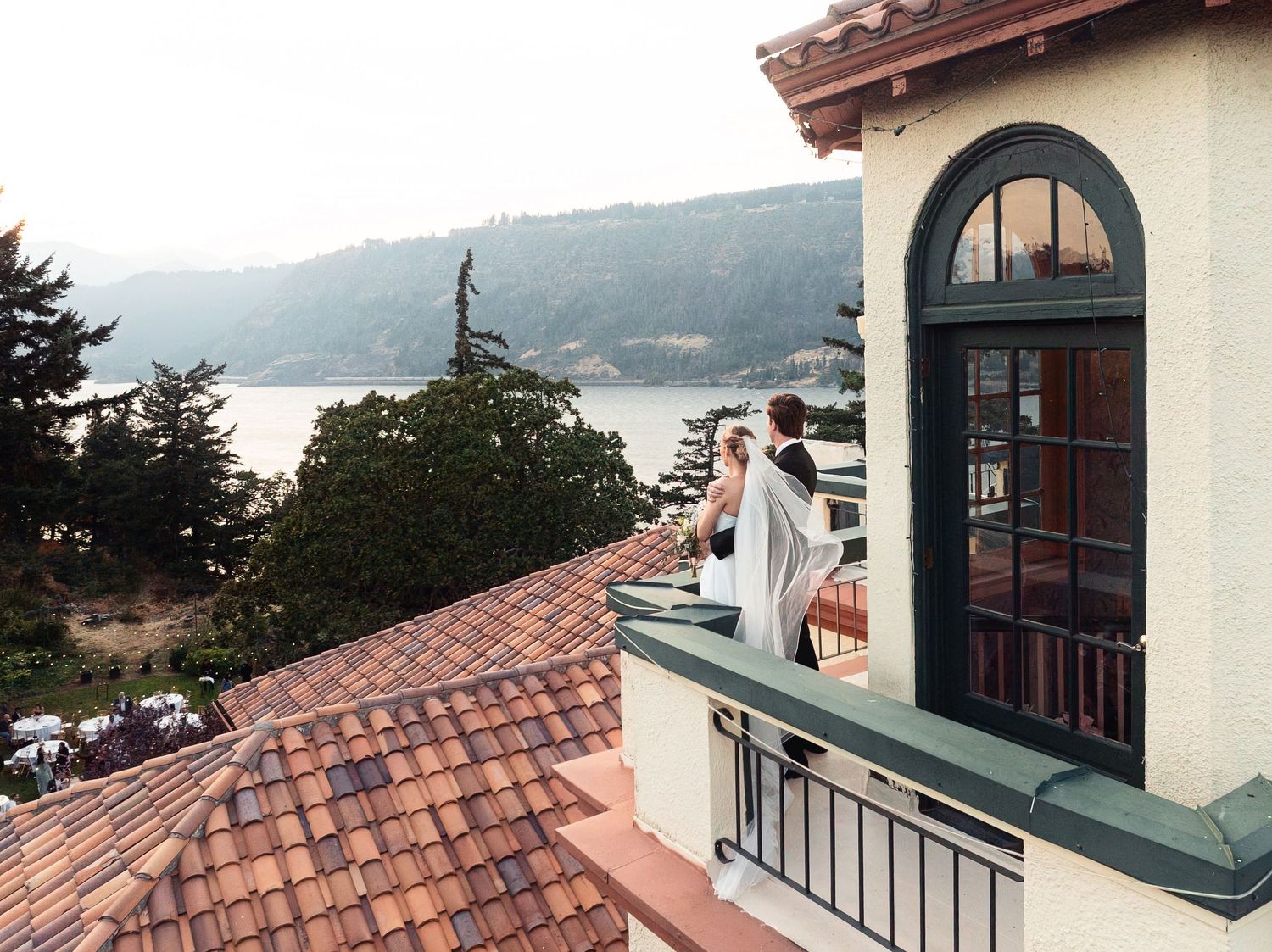 Bride and groom stand on a balcony overlooking a scenic bay with terracotta rooftops at a waterfront venue.