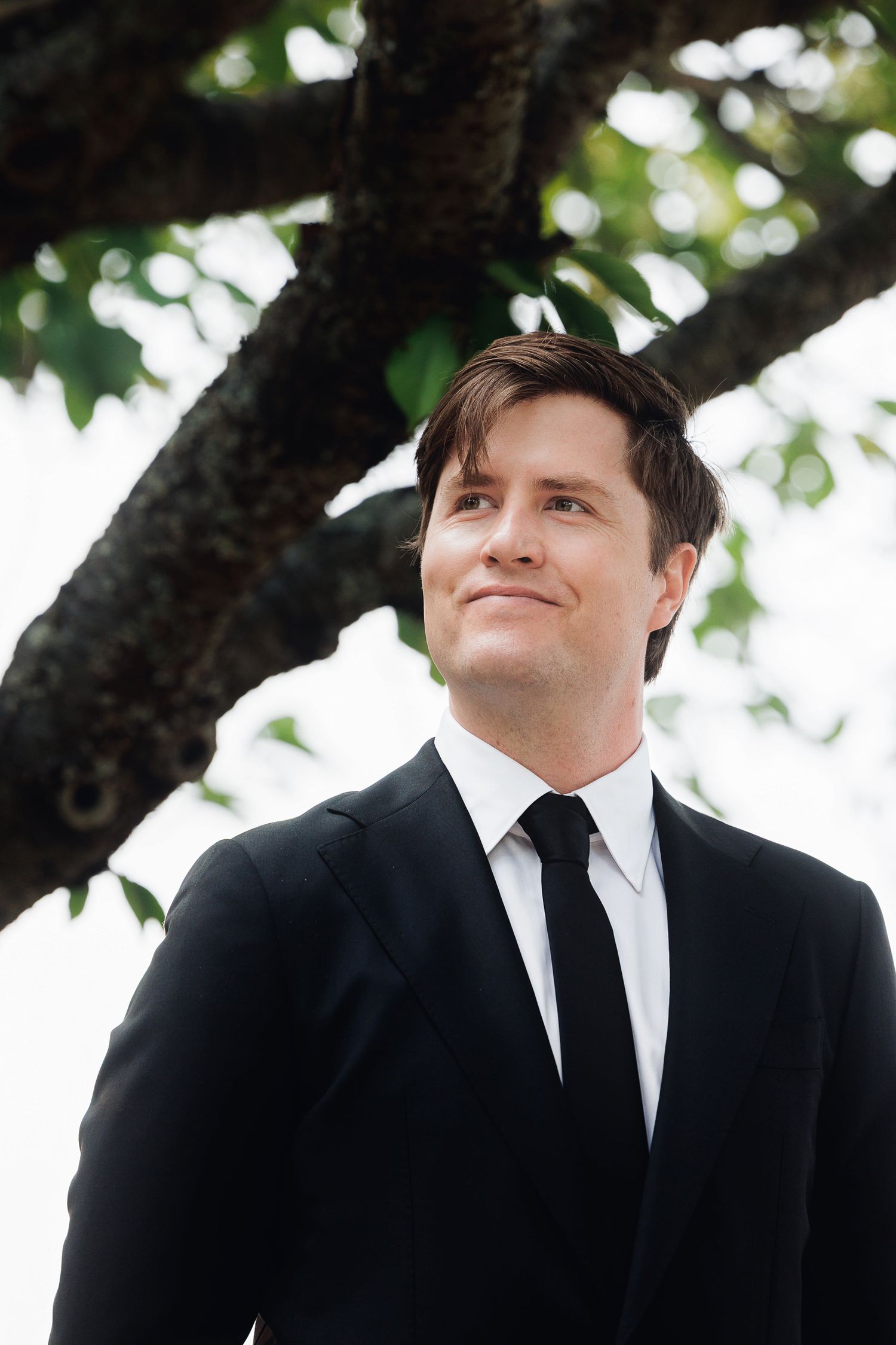 Man in black suit and tie smiling outdoors under a tree with green leaves in background.
