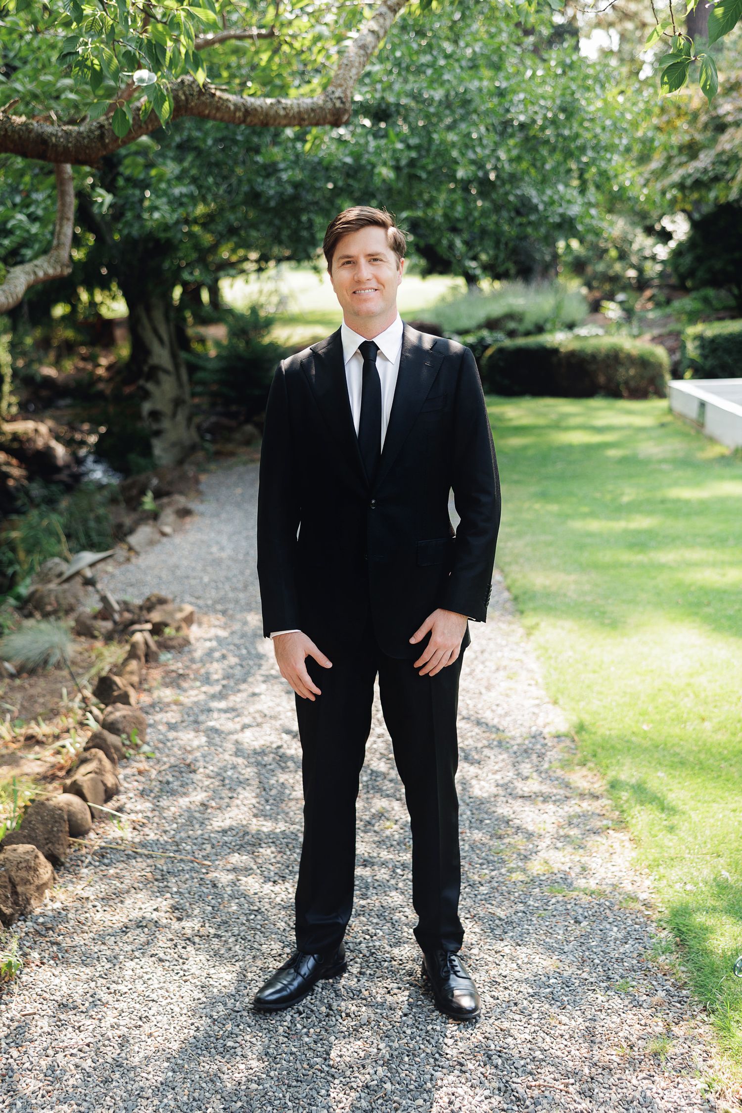 Groom in black suit and tie standing on gravel path surrounded by lush green garden trees.