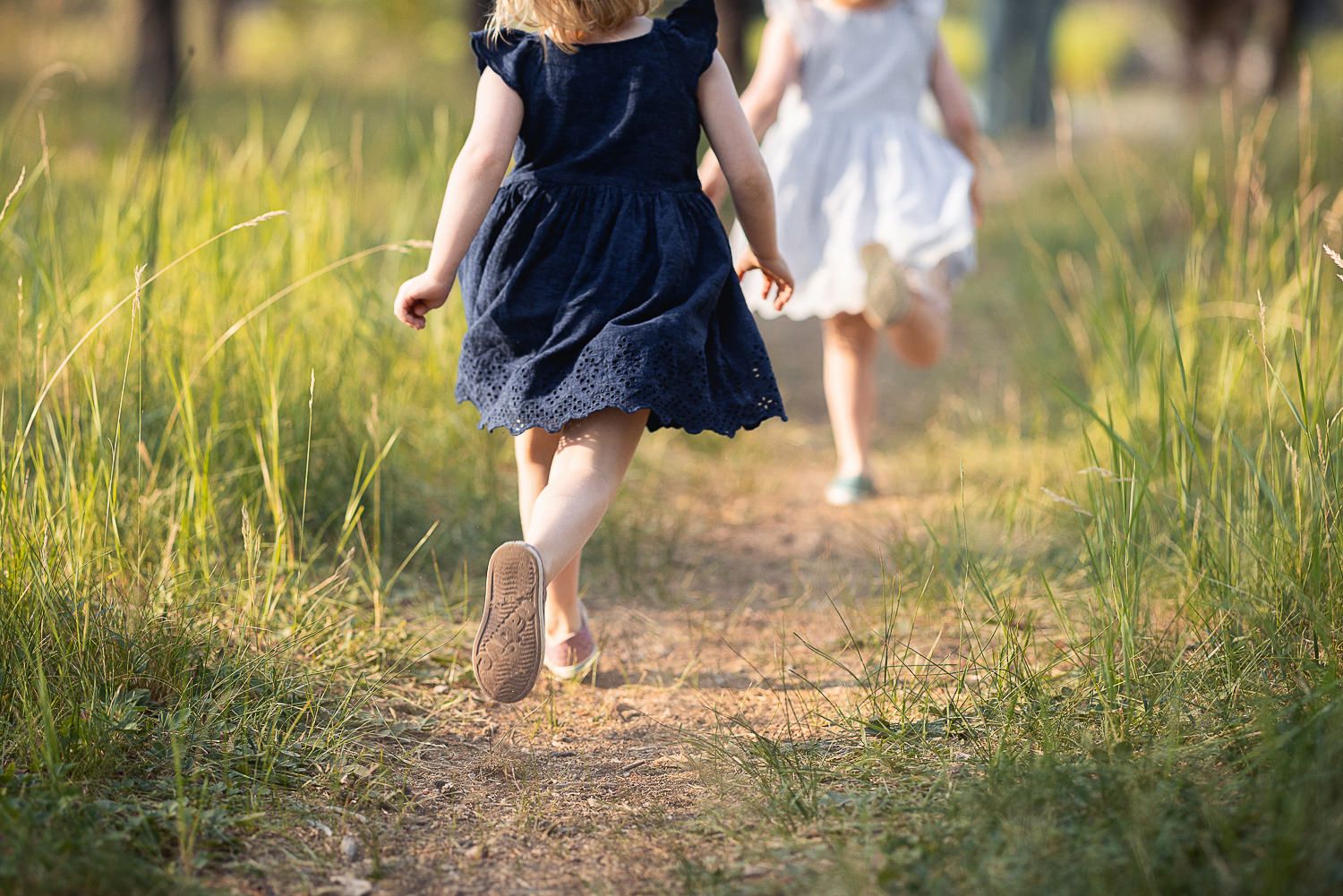 Two young children in dresses running barefoot on a dirt path through tall summer grass.