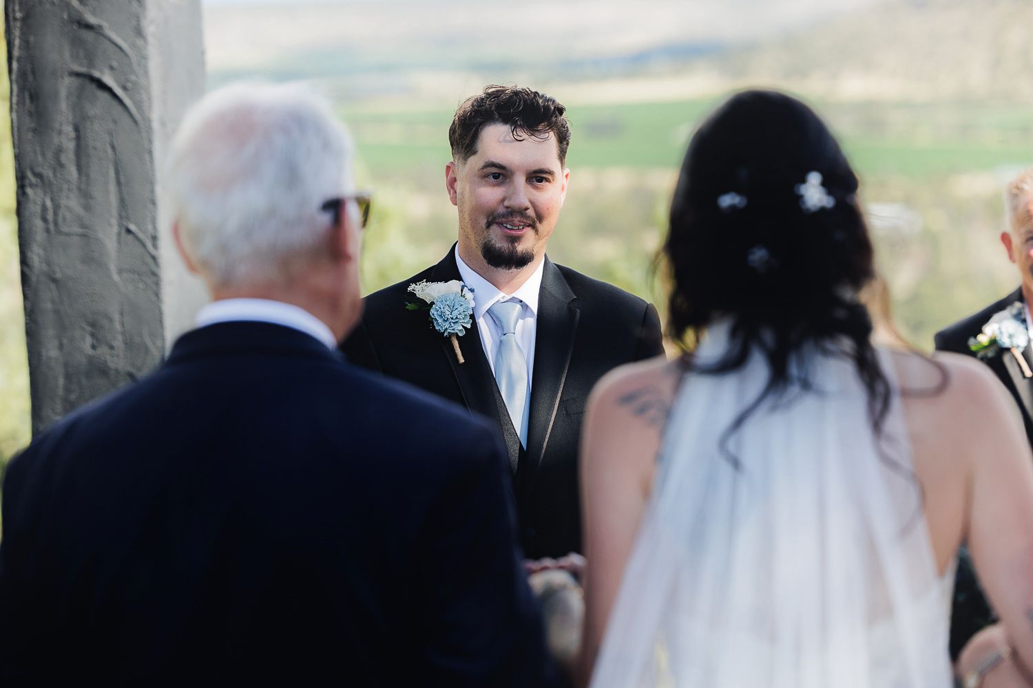 A wedding ceremony takes place outdoors with guests gathered around under natural lighting.