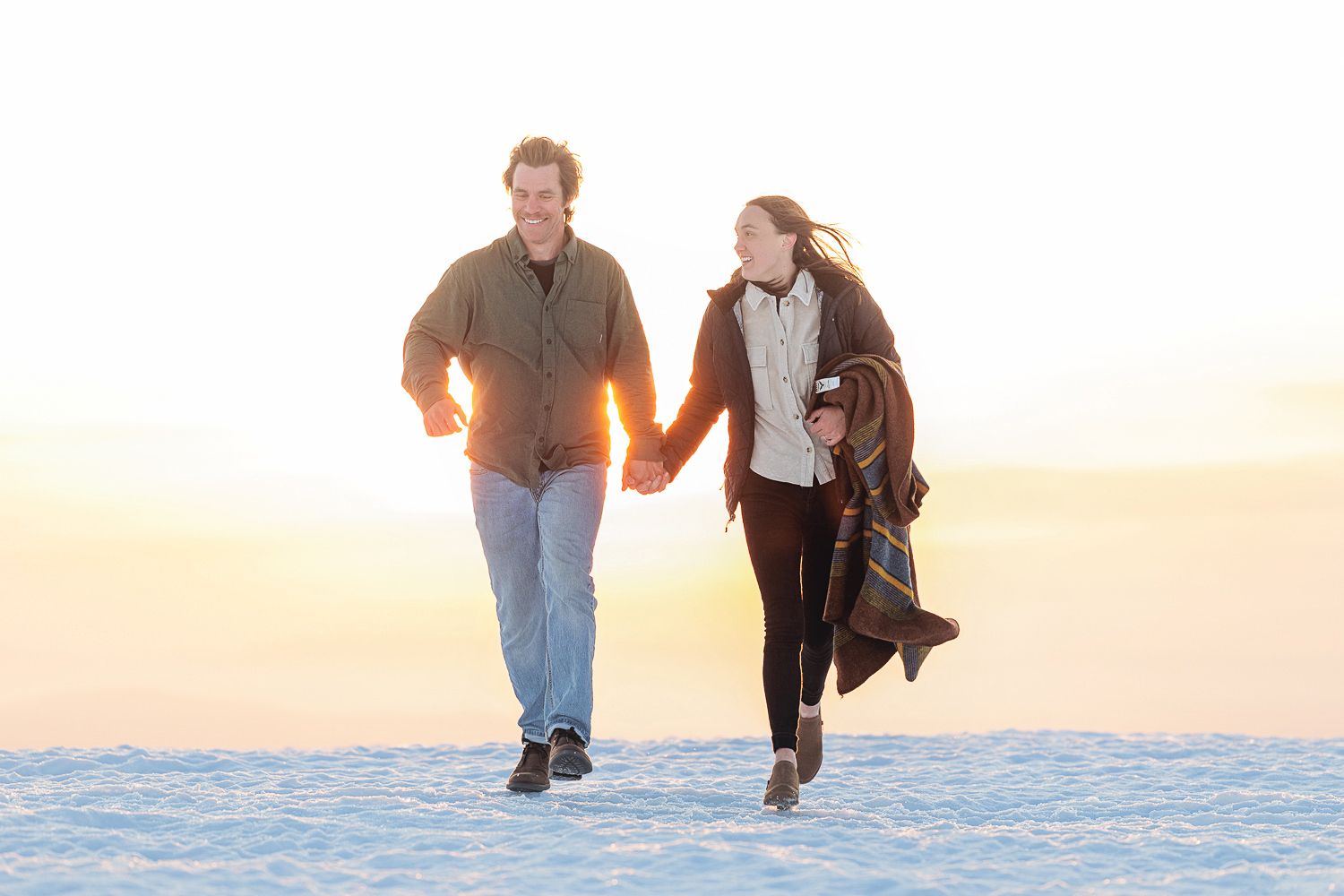 Couple holding hands while walking together on snowy ground during a golden sunset.