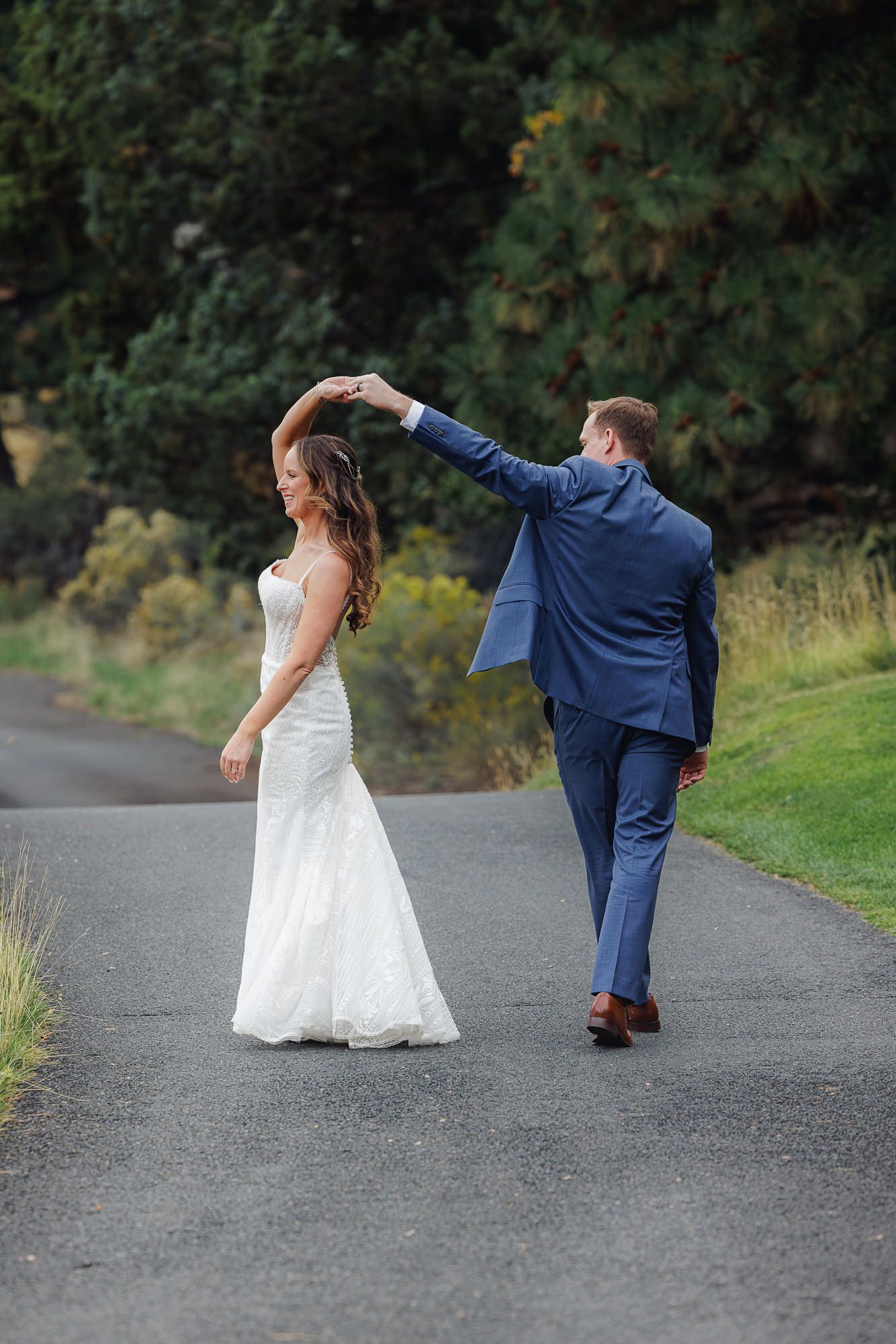 A couple in wedding attire dances and strikes playful poses on a paved path surrounded by greenery.