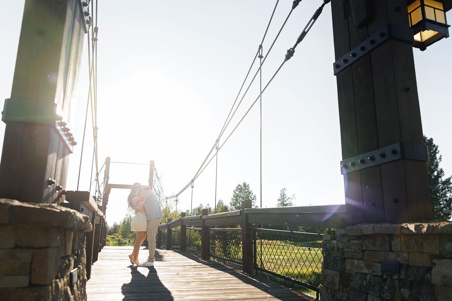 Person walking on a scenic wooden suspension bridge with stone towers on a sunny day.