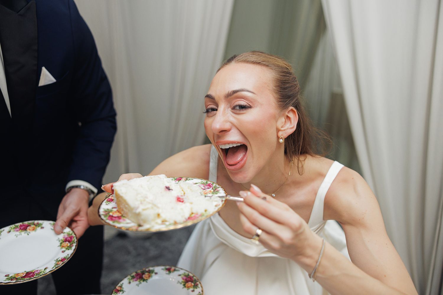 Excited bride laughing while eating white wedding cake at reception celebration.