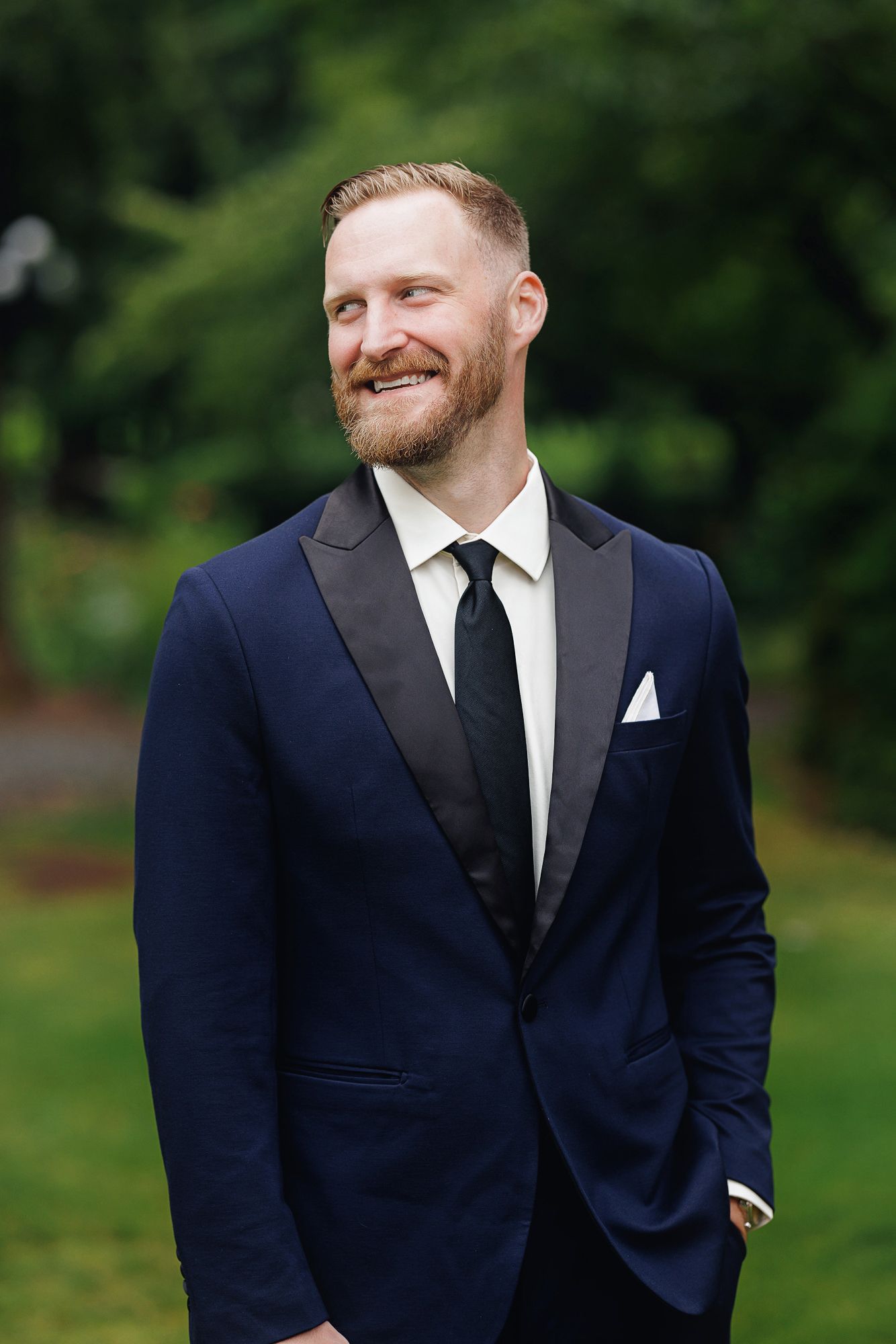 Smiling groom in navy tuxedo with black lapels and white pocket square outdoors.