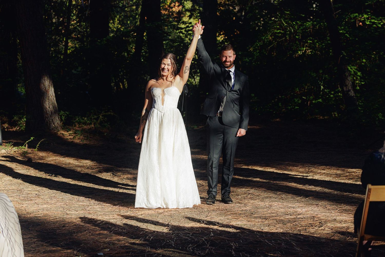 Bride and groom raising hands in celebration at outdoor forest wedding ceremony.