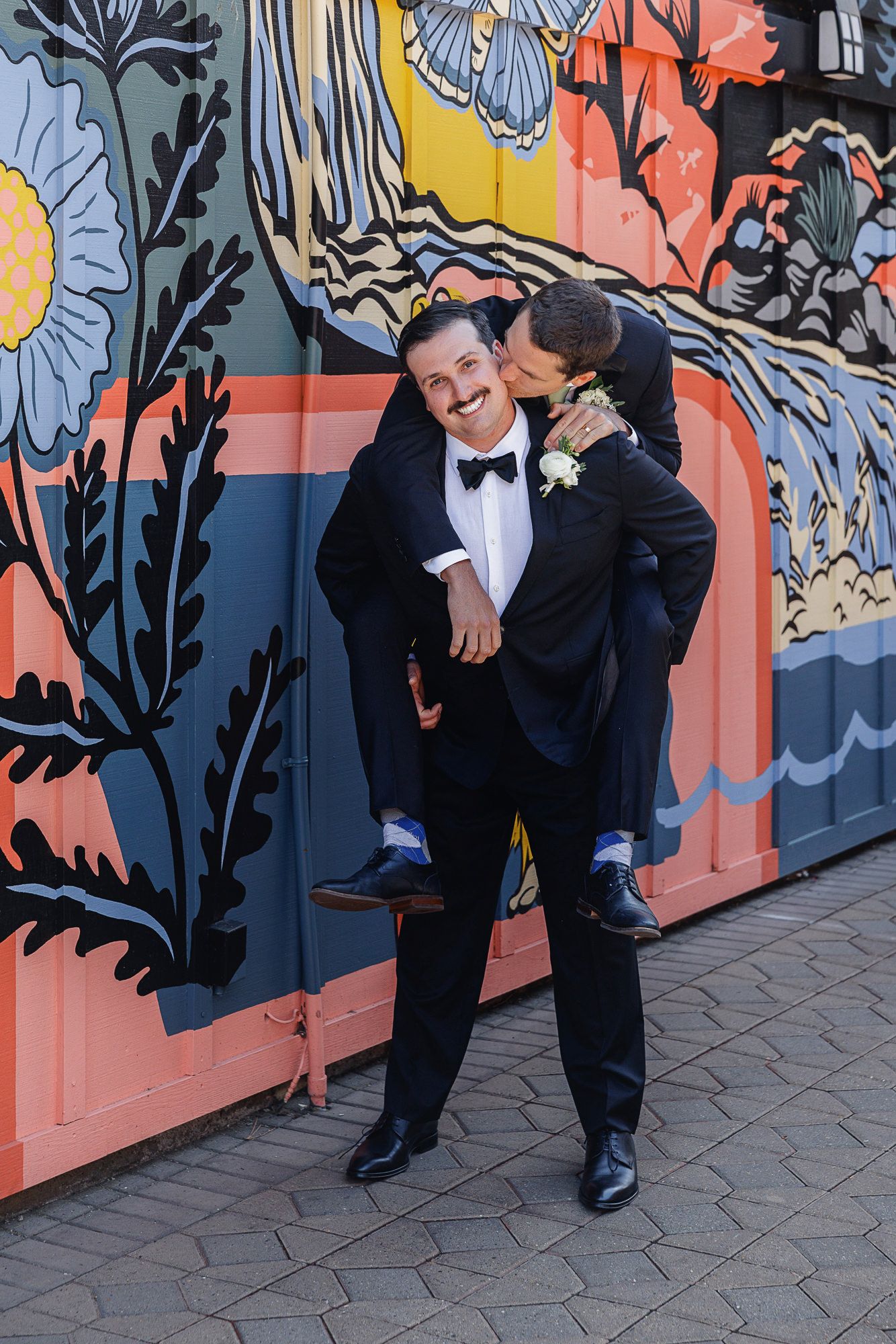 Two grooms in tuxedos pose playfully in front of a colorful floral mural on their wedding day.