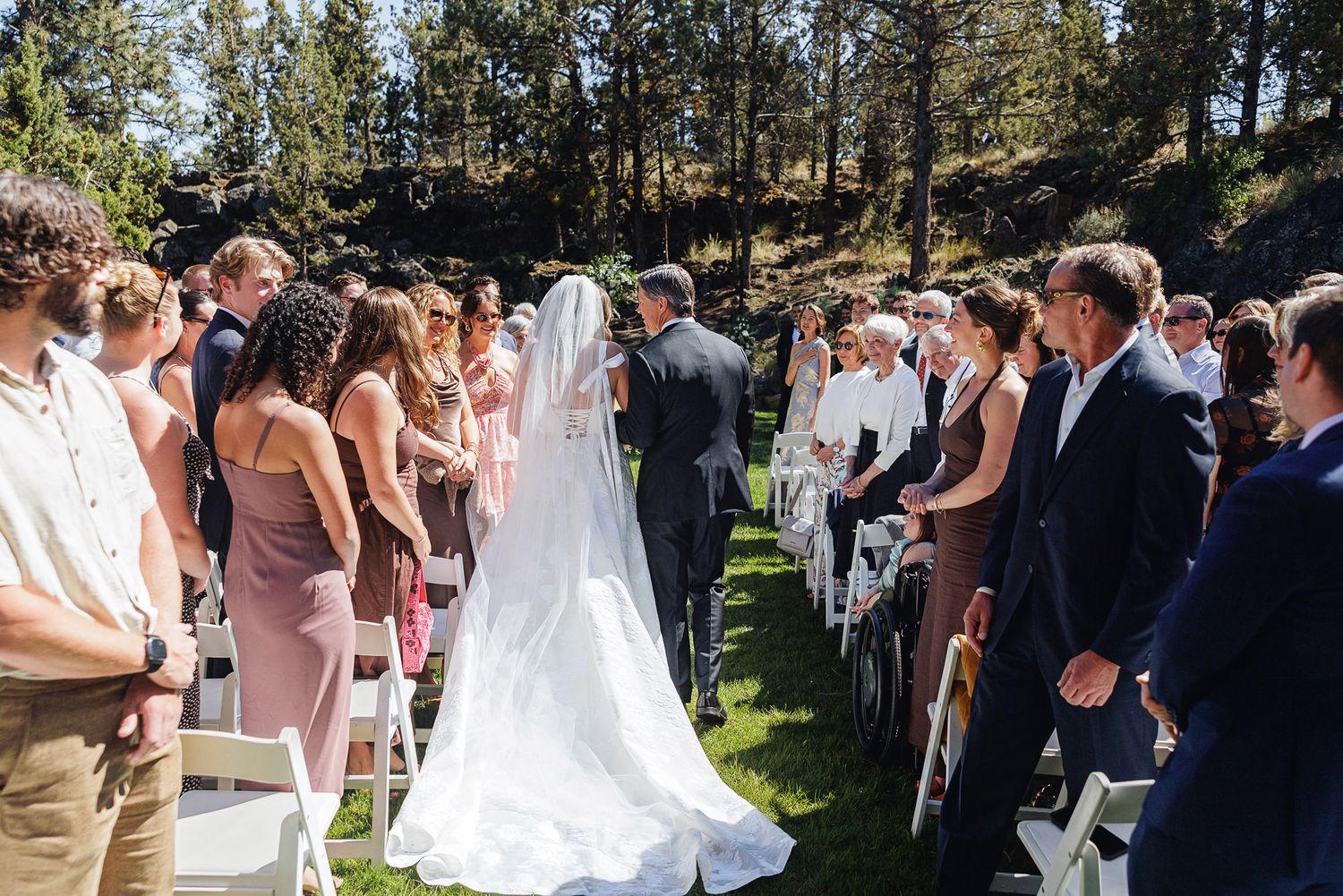 Bride in white gown and veil walks down the aisle with escort at outdoor wedding ceremony.