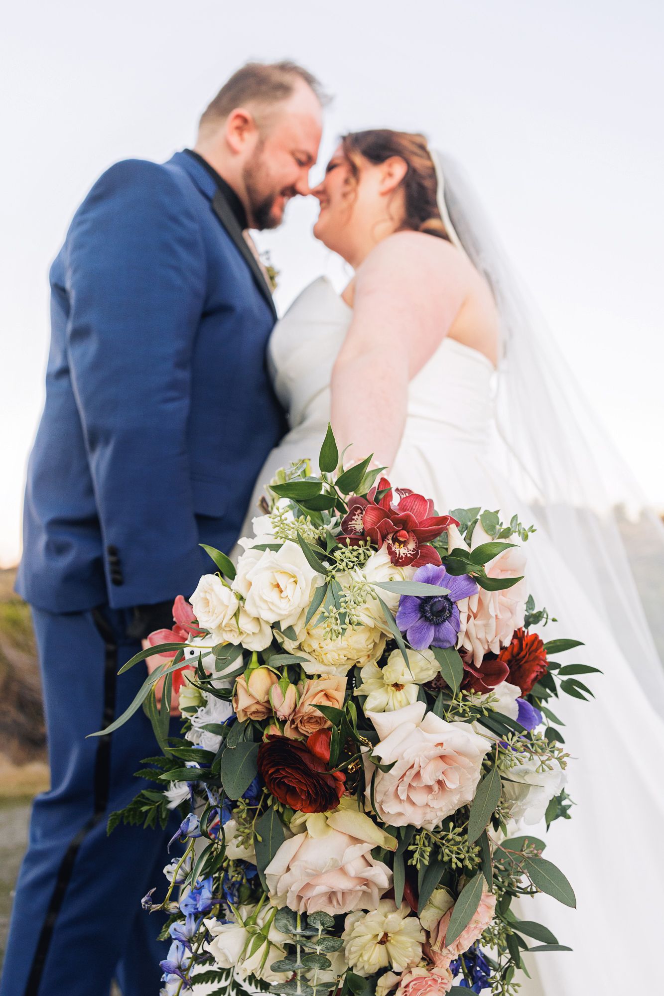 A romantic wedding scene with a navy-suited groom and bride holding a vibrant bouquet of mixed flowers in purple and white.